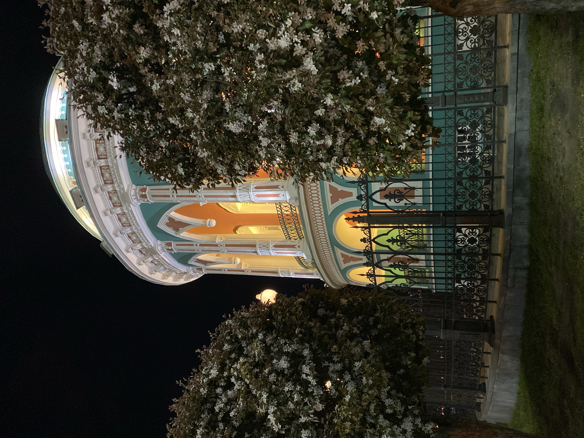 The image shows the exterior of the Kharitonov-Rastorguyev Mansion in Yekaterinburg, Russia, at night. The mansion features an ornate facade with arched windows illuminated from within. A decorative metal fence runs along the perimeter. Trees with white flowers partially obscure the view of the building. The sky is dark, indicating nighttime. The ground is a mix of grass and paving stones.