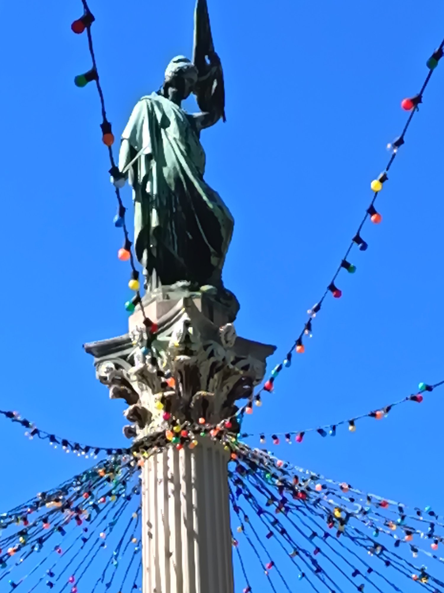 A low-angle view captures a statue atop a column, adorned with colorful string lights, set against a clear blue sky. The statue, positioned on an ornate capital, is draped in a flowing robe and holds an object aloft. The column is fluted and features decorative details at its base. The string lights, composed of bulbs in various colors, are draped extensively around the base of the column and hang down in strands. Some strands also extend upward, framing the statue. The location is the column of the Goddess in Nancy, France.