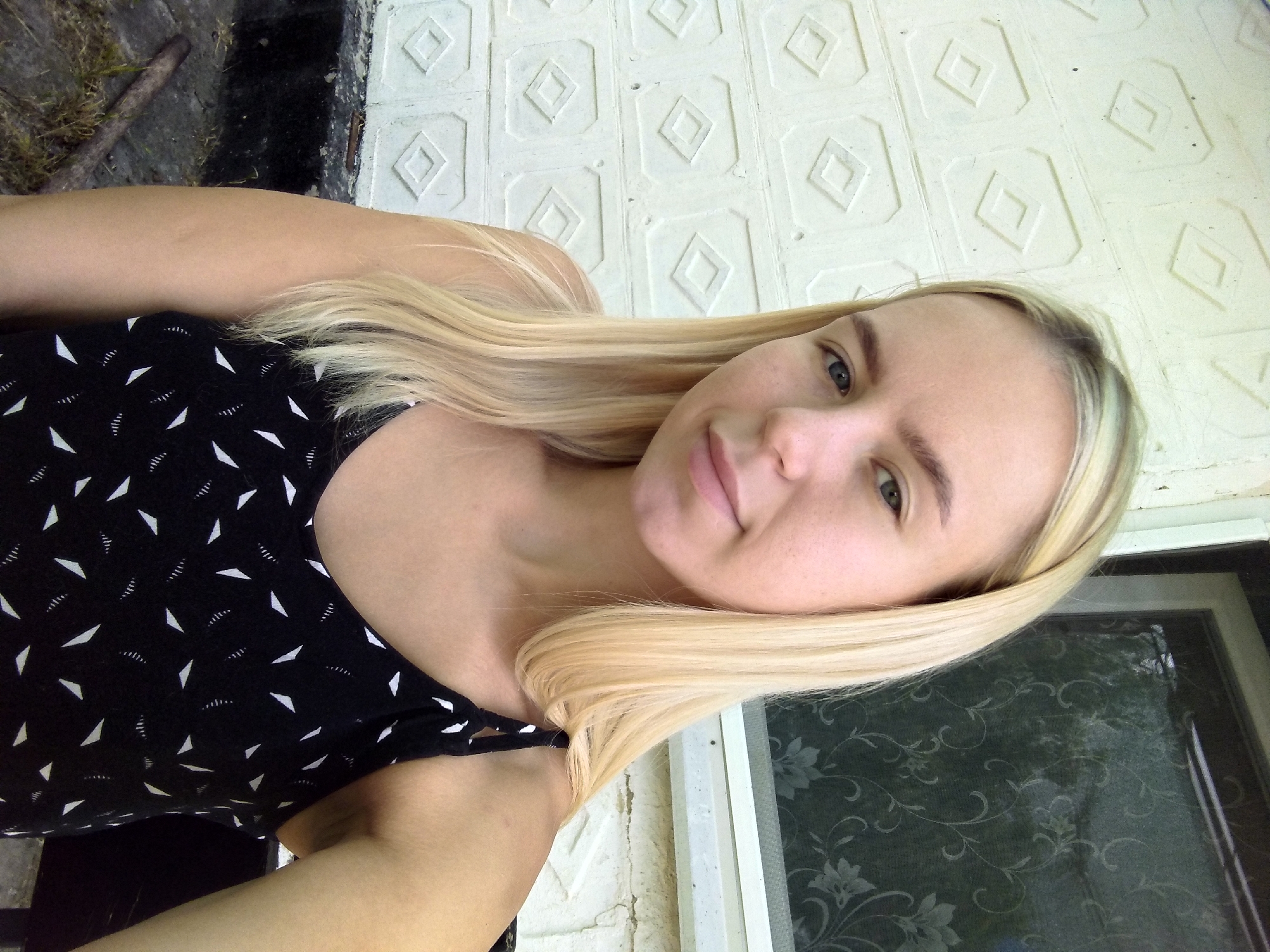A young woman with blonde hair and blue eyes, wearing a black tank top with a white geometric pattern, poses for a selfie outdoors. She is positioned in front of a textured white wall with diamond-shaped insets and to the right of a window with a floral screen. The environment suggests a residential area, possibly in the city of Radisne, Ukraine, given the architectural style. The ground in the foreground consists of concrete and dry grass.