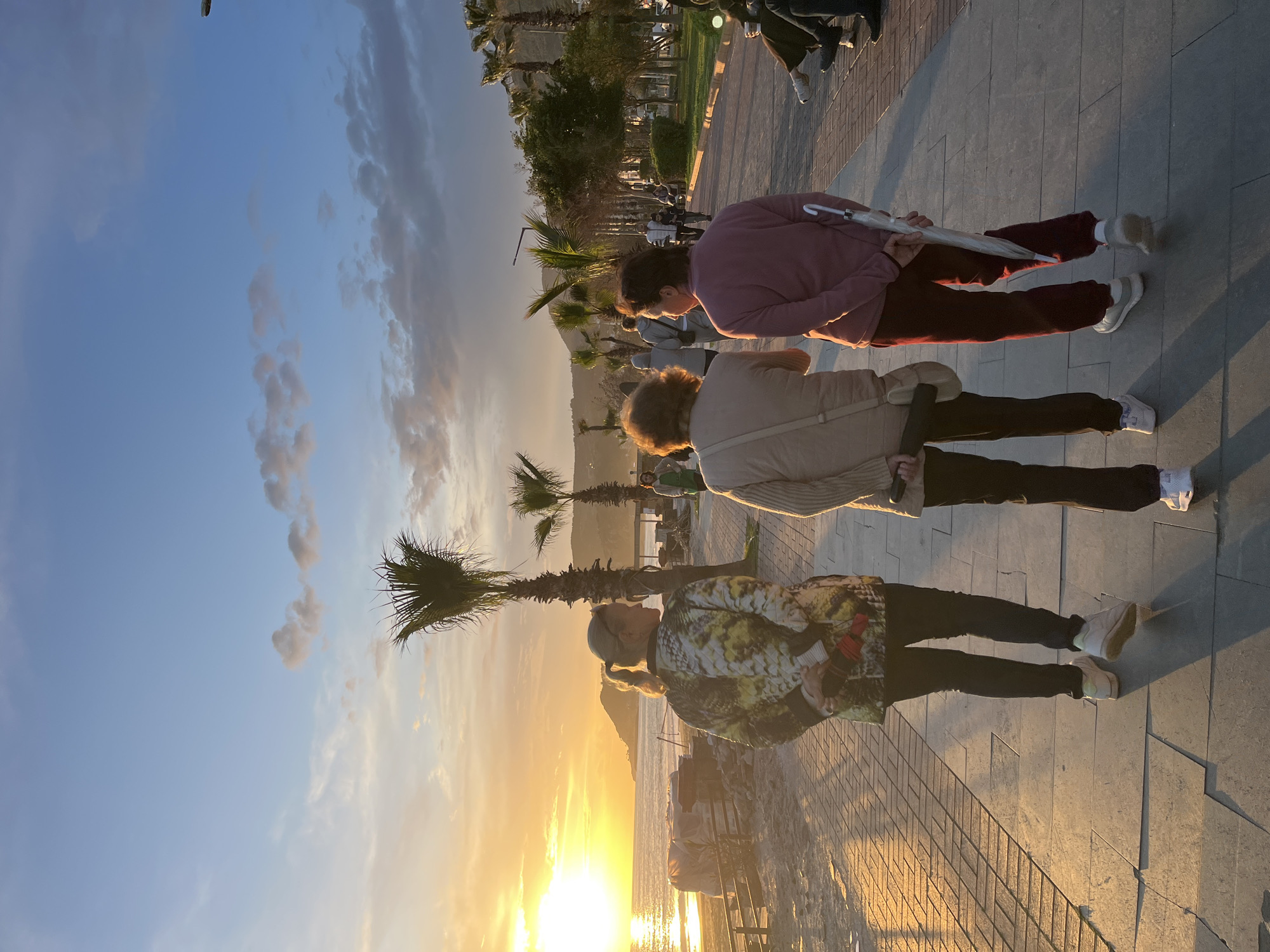 The image captures a scene in Alanya, Turkey at sunset. Three women are standing on a paved walkway, likely a promenade. The sun is setting over the water, casting a golden glow on the scene. The woman closest to the viewer is wearing a patterned jacket and dark pants. The second woman is wearing a beige jacket and dark pants, and the third woman is wearing a pinkish-red shirt and maroon pants, holding a silver umbrella. Palm trees are visible along the walkway. Other people can be seen in the background, further along the promenade. The sky is a gradient of blue and orange, with some clouds present. The sea reflects the light of the setting sun.