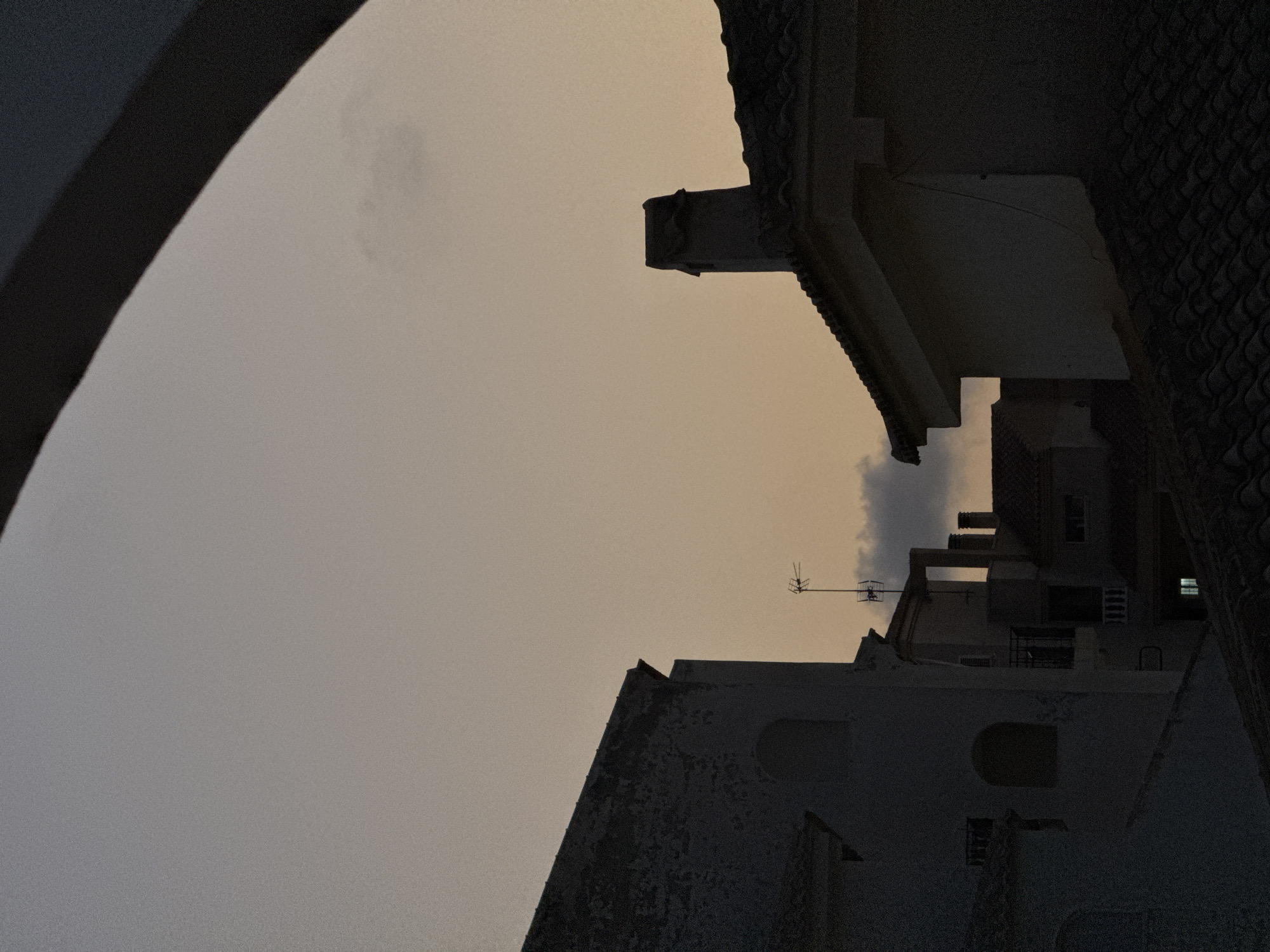 The image depicts an exterior view of rooftops and buildings in Torrevieja, Spain, during what appears to be dusk or dawn. The sky is a muted, pale orange-grey with a few wispy clouds visible. In the foreground, a dark, curved architectural element frames the top of the scene. Silhouetted against the sky are the roofs and upper levels of several buildings. On the left, a building with a textured, light-colored facade features an arched window opening. To the right, another building has a dark, tiled roof and a prominent, rectangular chimney. In the mid-ground, other buildings are visible, including one with an antenna. Some windows are illuminated, suggesting occupancy. The overall impression is one of quiet, urban twilight.