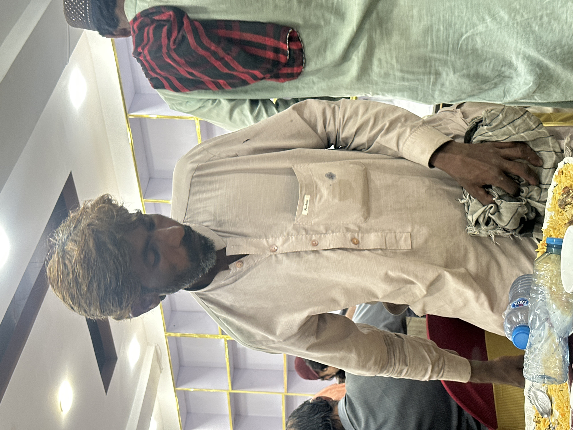 A medium shot depicts a man with graying hair and a dark beard wearing a light beige button-down shirt. He is holding a gray and white checkered cloth in his right hand. To the right, there are clear plastic water bottles and a plate of food, possibly rice. Behind him and slightly above, a man wears a green garment with a red and black plaid scarf around his neck. The background shows a pale wall with gold trim or shelving. The scene appears to be indoors, likely in the city of Quetta City Tehsil, Pakistan.