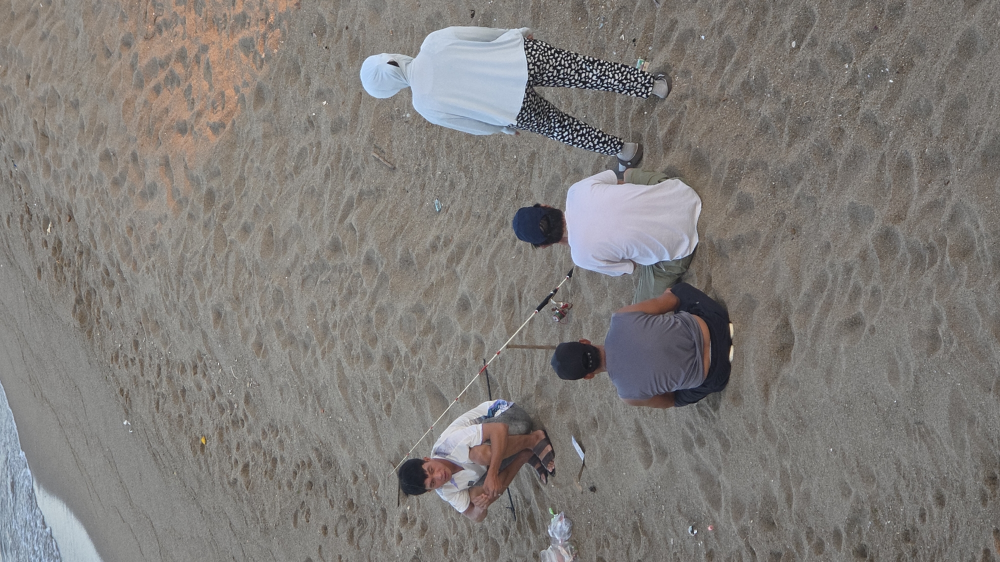 The image displays a sandy beach in Nha Trang, Vietnam. Four individuals are present on the light brown sand, which shows numerous footprints and scattered small debris, including plastic items and sticks. Gentle ocean waves are visible along the upper left edge of the frame.
On the left, an Asian male squats on the sand, facing slightly right and forward. He wears a white short-sleeved shirt with blue patterns, dark shorts, and dark sandals, with his hands clasped between his knees. Adjacent to him are a clear plastic bag, an open plastic container, and a metallic object on the sand. A fishing rod, propped on a stick, extends to his right with a red reel visible.
To the right of the squatting individual, two Asian males are seated on the sand with their backs to the camera. The man closer to the fishing rod wears a white short-sleeved t-shirt, light pants, and a dark baseball cap. The man to his left wears a dark grey sleeveless shirt, dark pants, and a dark baseball cap worn backwards.
Further to the right, an adult figure stands with their back to the camera, wearing a light blue long-sleeved top that includes a head covering, black and white patterned trousers, and light-colored shoes. The sun casts a warm, orange hue on sections of the sand.