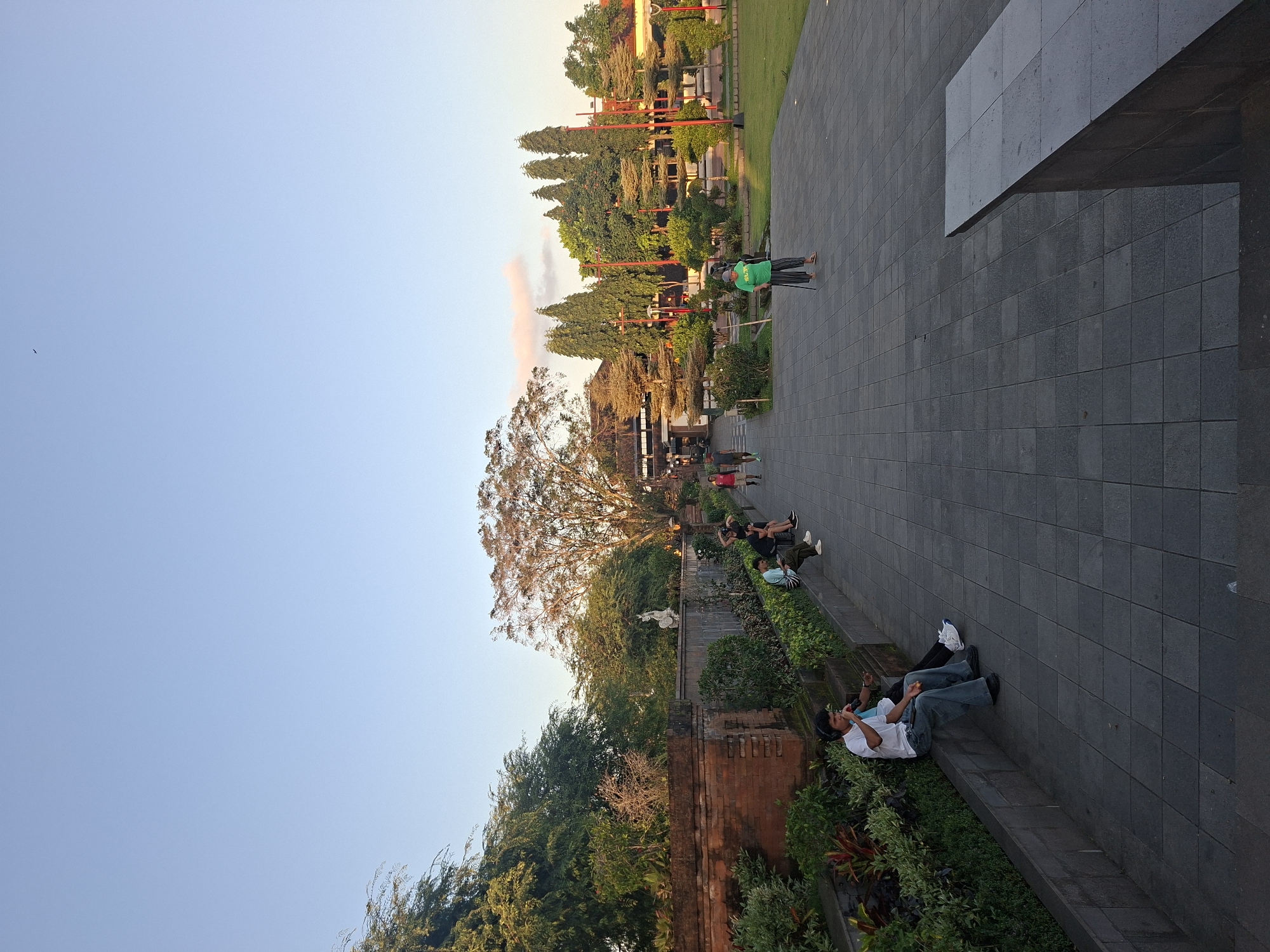 The image depicts an outdoor scene in the city of Gianyar, Indonesia, likely during late afternoon or early evening based on the lighting. The foreground features a wide paved pathway with a textured, tiled surface. To the left, a low, brick wall is covered with lush green plants and colorful foliage, suggesting a garden or manicured landscape. Several individuals are seated on the edge of this wall, some interacting or resting. One man in a white t-shirt and jeans is holding a microphone and wearing headphones, possibly performing or engaged in a recording.
Further back, the pathway leads towards a large, traditional-looking building with dark wood and tiered roofing, suggesting it could be a temple or cultural site, characteristic of Balinese architecture. The area around the building is a grassy park with numerous trees, including tall, slender ones and broader, leafy trees. Several people are visible walking or standing in this area. One man in a green shirt is standing with a cane, looking towards the building. The sky is clear and light blue with a hint of pinkish clouds near the horizon. A single bird is visible in flight against the vast expanse of the sky. The overall impression is a public space, possibly a tourist attraction or a gathering place, with a blend of architectural elements and natural greenery.