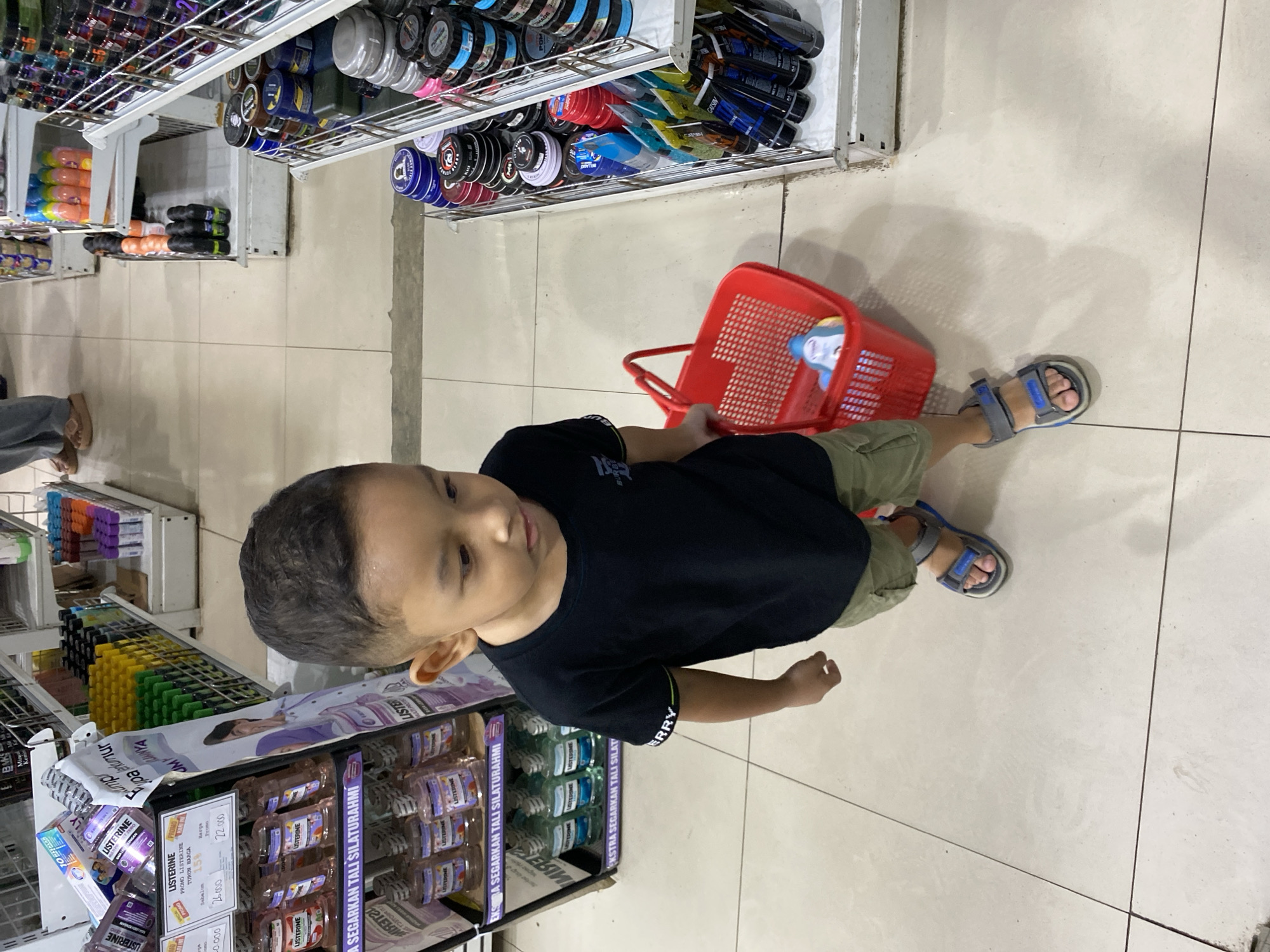 In a retail store in Lampisang, Indonesia, a young boy stands in the foreground, looking down and to his right. He is wearing a black t-shirt and khaki shorts, with blue and gray sandals on his feet. In his right hand, he holds a red plastic shopping basket containing a single blue and white item, possibly a bottle or toy. The boy is positioned on a light-colored tiled floor. Behind him, to his left, is a display rack filled with various bottles of Listerine mouthwash and other health and beauty products. To his right, another display shelf is stocked with numerous jars and tubes of hair styling products. In the background, other shoppers and store aisles are visible, suggesting a typical busy retail environment. The overall scene depicts a child shopping with his family in a well-lit store.