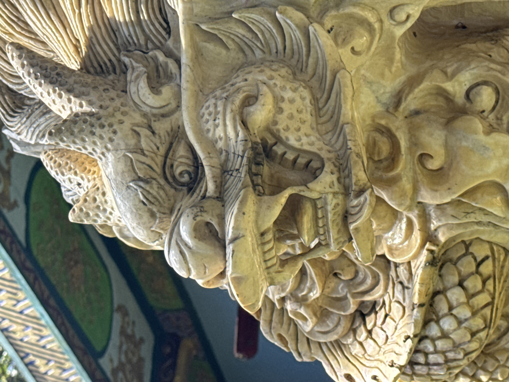 The image is a close-up, detailed shot of a carved stone dragon sculpture, likely an architectural element or a statue, positioned outdoors. The dragon's head is prominently featured, with its mouth open revealing sharp teeth and a detailed, textured tongue. Its scales and mane are intricately carved, giving the impression of movement and power. The sculpture appears to be made of a light-colored stone, possibly marble or a similar material, with visible weathering and some darker lines suggesting age or dirt. In the blurred background, a portion of a building is visible, featuring decorative patterns in blue, green, and gold, typical of traditional Asian architecture. A red horizontal beam is also visible in the background. The lighting suggests it is daytime, with sunlight illuminating the sculpture from the side, casting subtle shadows that enhance the carving's dimensionality. The sculpture is located in Xuân Thành, Vietnam, likely a historical or cultural site given the architectural style and decorative elements in the background.