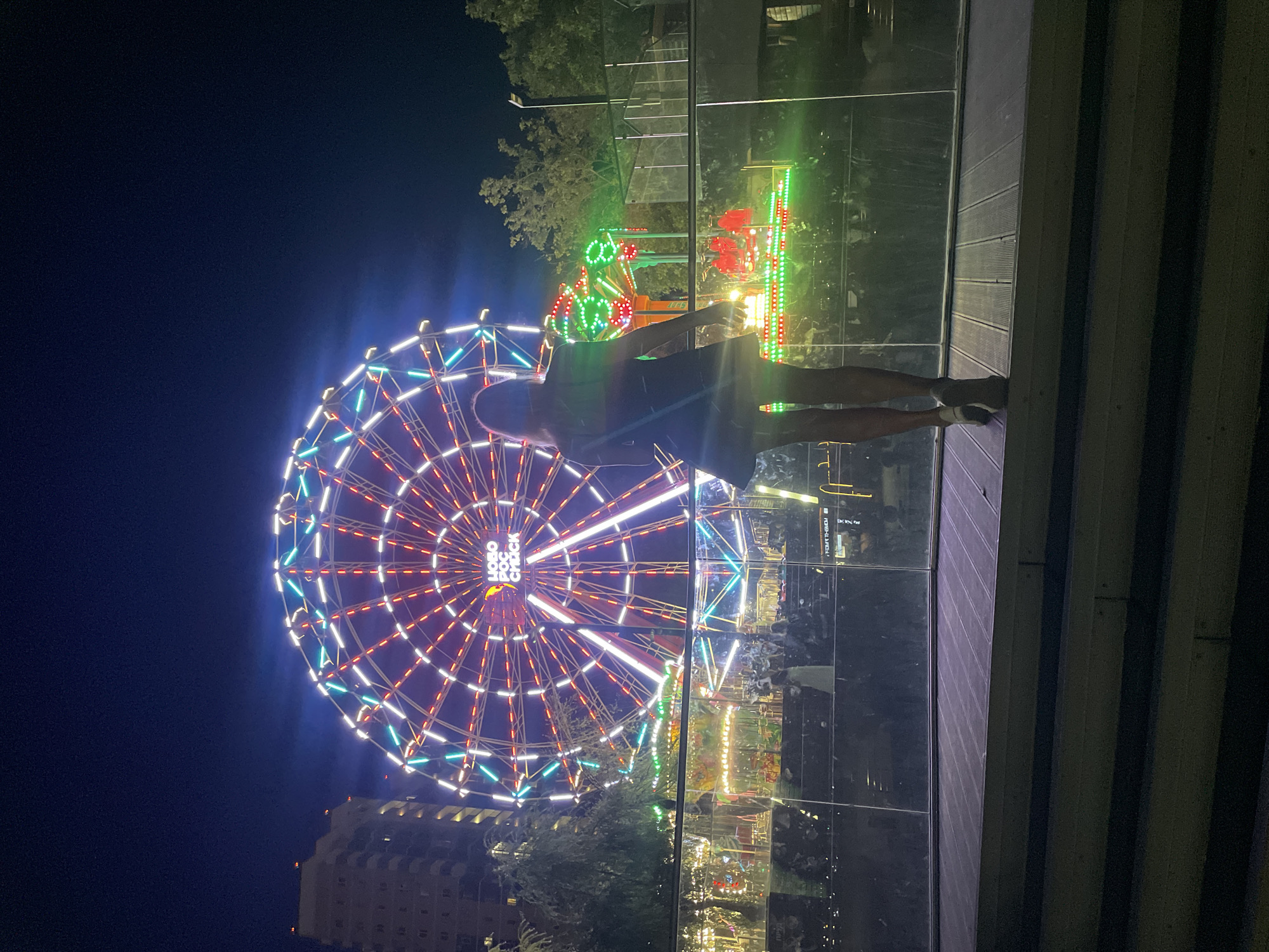 The image shows a Ferris wheel illuminated with colorful lights at night, located in the city of Novorossiysk, Russia. A woman, viewed from the back, stands on a wooden platform in the foreground, looking towards the Ferris wheel and other amusement park attractions. The Ferris wheel is a large, circular structure with bright red, white, and blue lights. Below the Ferris wheel, there are smaller amusement rides with green and red lights. A tall building is visible to the left of the Ferris wheel. The scene is set at night, with the sky appearing dark blue. The woman is wearing a short, dark dress and high heels. She is standing with her arms at her sides. The wooden platform has a textured surface. A glass railing is positioned between the platform and the amusement park attractions.