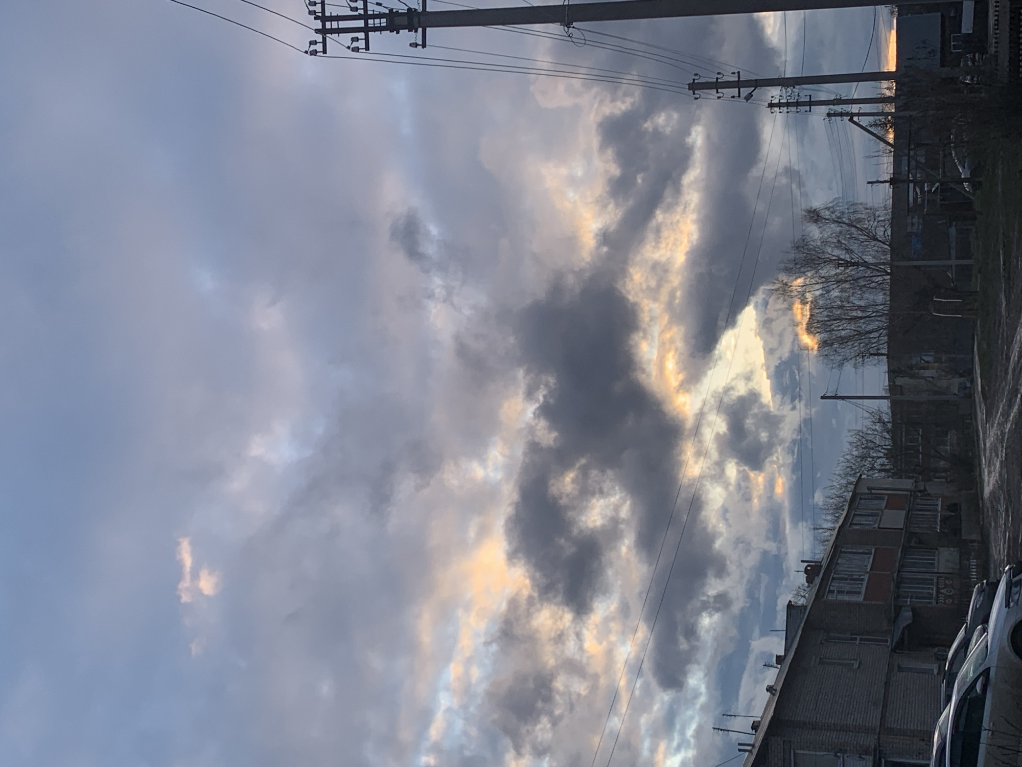 The image displays a dramatic cloudy sky during what appears to be either dusk or dawn, with warm, golden light piercing through heavy, dark grey cloud formations. The sky dominates the composition, featuring patches of pale blue interspersed with the clouds.
Prominently featured in the upper and middle sections are multiple utility poles and a dense network of overhead power lines, silhouetted against the sky. These structures stretch across the frame, adding a linear element to the scene.
Below the horizon line, to the right and bottom right, are parts of multi-story buildings, likely residential, with visible windows and a somewhat weathered appearance. Leafless trees, suggesting winter or early spring, stand in front of some of these structures, their branches silhouetted. On the far bottom right, the tops of a couple of parked cars are visible.
The scene is static, with no discernible human activity. The overall mood is atmospheric, characterized by the interplay of light and shadow from the heavy cloud cover. The environment suggests an urban or suburban setting, consistent with a location like Фоки, Russia. There is no visible text in the image.