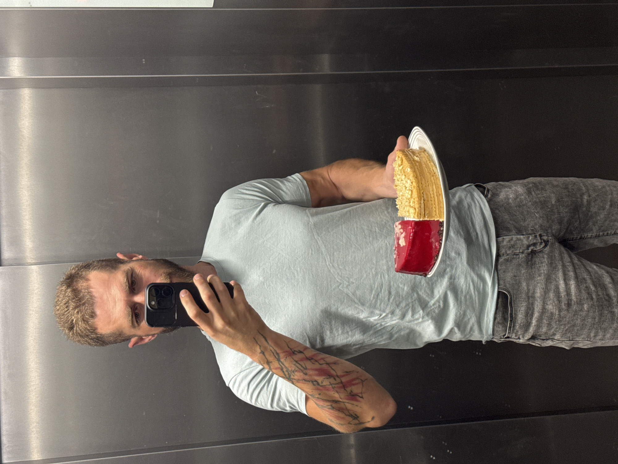 A man with short, blond hair and a beard is standing in an elevator and taking a mirror selfie with his phone. He is wearing a light blue t-shirt and grey jeans. He is holding a plate with a half-red and half-yellow cake. The elevator has brushed metal walls. There is no indication of the city of Budva, Montenegro.