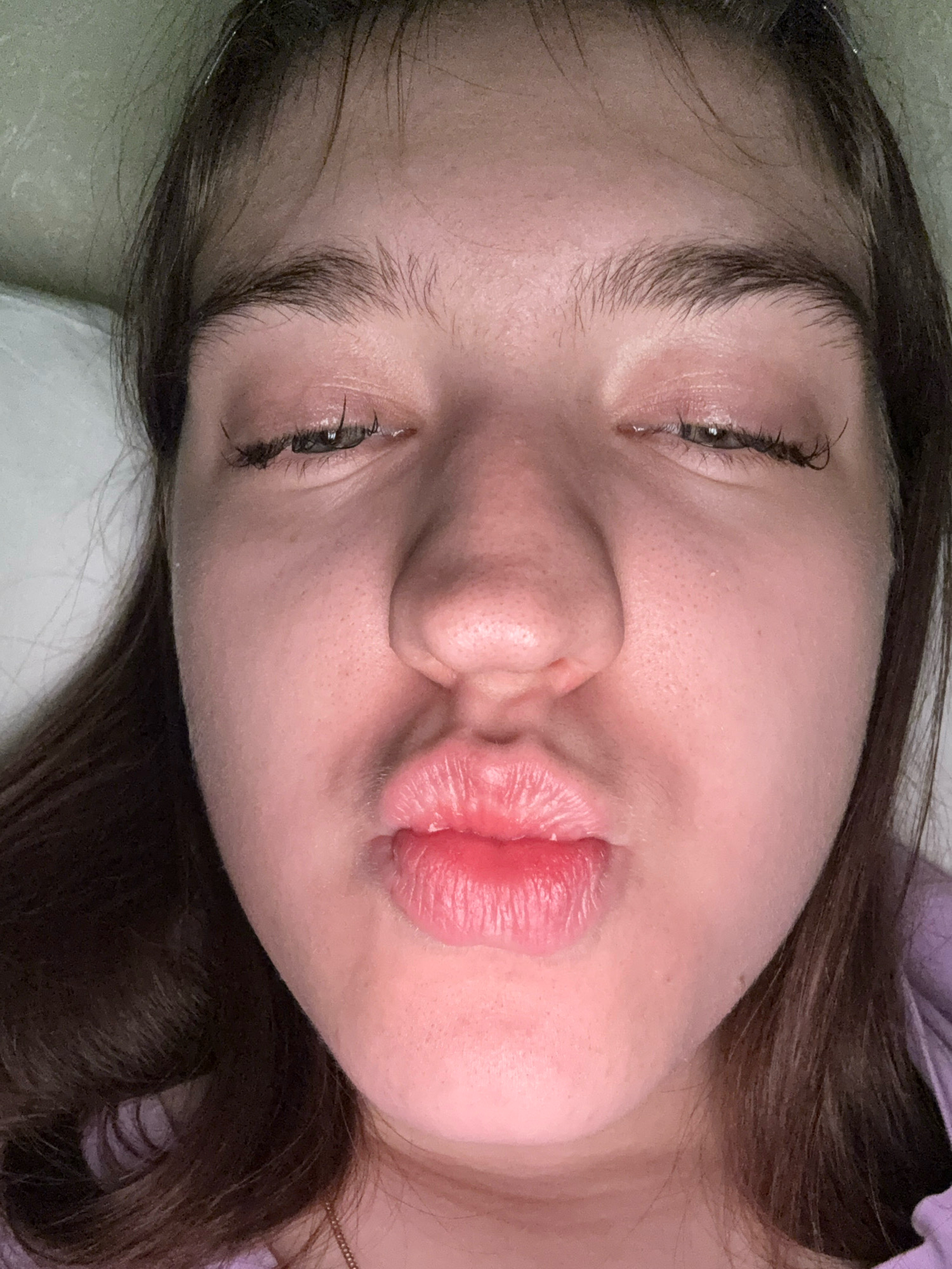 A young woman with brown hair is shown in a close-up selfie, with her eyes partially closed and her lips puckered as if for a kiss. Her skin is pale and appears to have some texture, with visible pores. She is wearing a light purple top. The background is indistinct and appears to be indoors, possibly a bed with a white pillow. There is no information available to place this image in Zavolzhye, Russia.