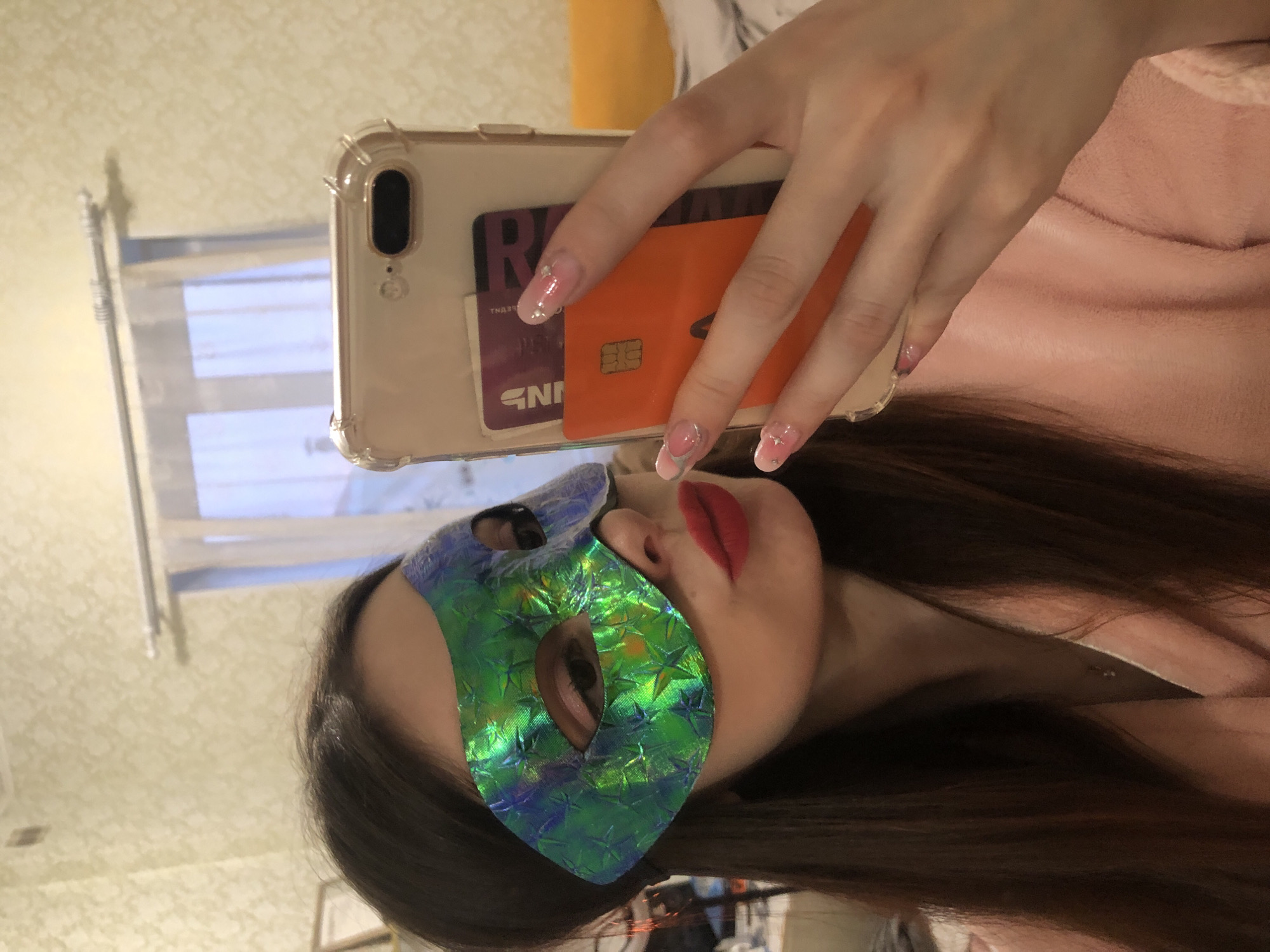 A portrait-oriented image shows a woman taking a selfie, likely reflected in a mirror. The woman's face is partially obscured by a vibrant green and blue iridescent mask with a star pattern, revealing only her eyes and red-lipsticked lips. Her long, dark hair frames her face.
Her right hand holds a smartphone with a clear protective case. Tucked into the back of the phone case are two cards: one purple with partially visible text like "R" and "NN" (possibly a loyalty or bank card), and one orange, which appears to be a chip card. Her fingernails are painted with a pink and white French manicure style, adorned with tiny star or glitter details. She is wearing a soft, pink garment, possibly a robe or sweater.
In the background, a patterned light-colored wallpaper is visible, along with a window and a curtain rod, suggesting an indoor setting. The scene conveys a playful or mysterious mood due to the mask. This image was captured in Noviy, Russia.