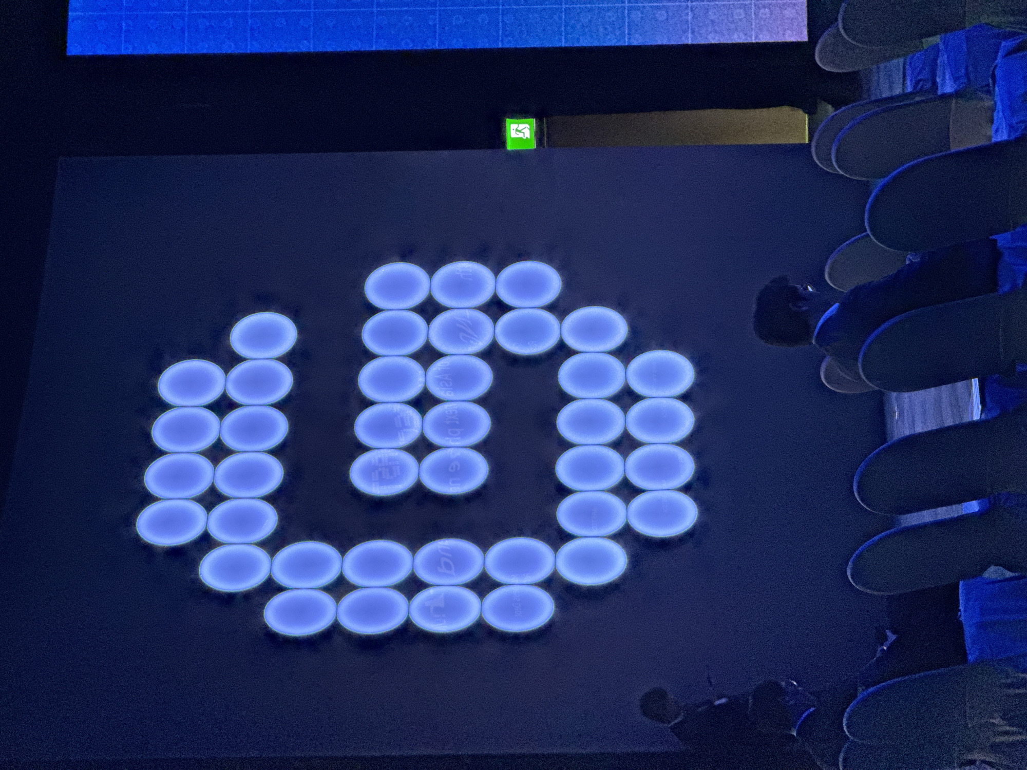 The image depicts a dark conference room with a large, illuminated "G" logo made of blue circles on a screen. The logo is the primary subject, taking up most of the frame. Below the screen, the backs of several chairs are visible, some occupied by individuals. The lighting is dim, with the blue glow from the screen casting a blue hue on the room. The scene appears to be a presentation or event. There is an illuminated green exit sign on the right side of the screen.