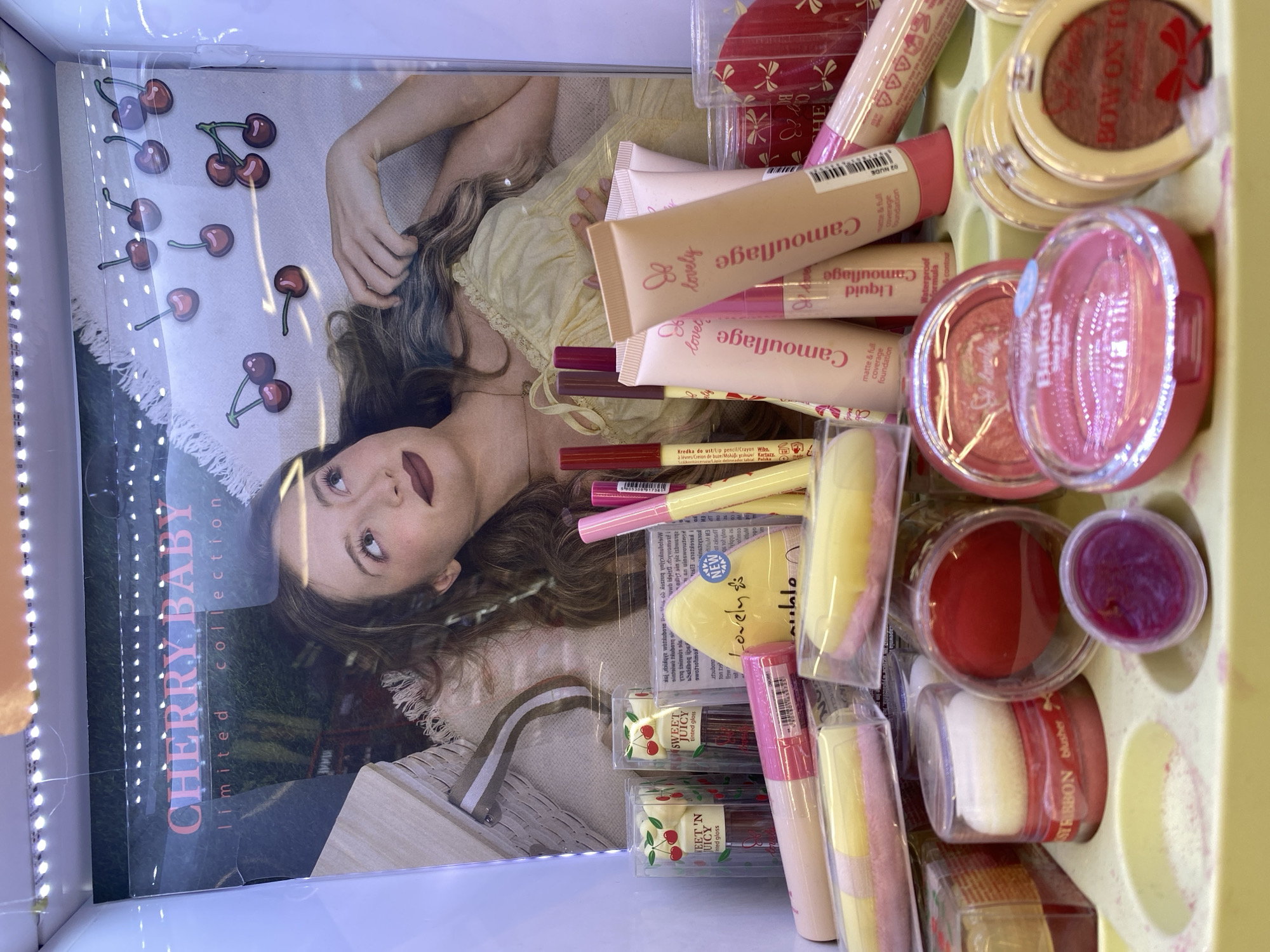 The image shows a display of makeup products, primarily from the "Lovely" brand, in what appears to be a retail setting. The backdrop features a poster titled "CHERRY BABY limited collection" with an image of a young woman and cherries. The foreground displays various cosmetic items such as lip glosses, liquid foundations, blushes, and makeup sponges, all arranged on a yellow molded plastic tray. The lighting suggests an indoor, well-lit environment, possibly a store shelf or counter. There is no discernible location in the city of Buk, Poland.