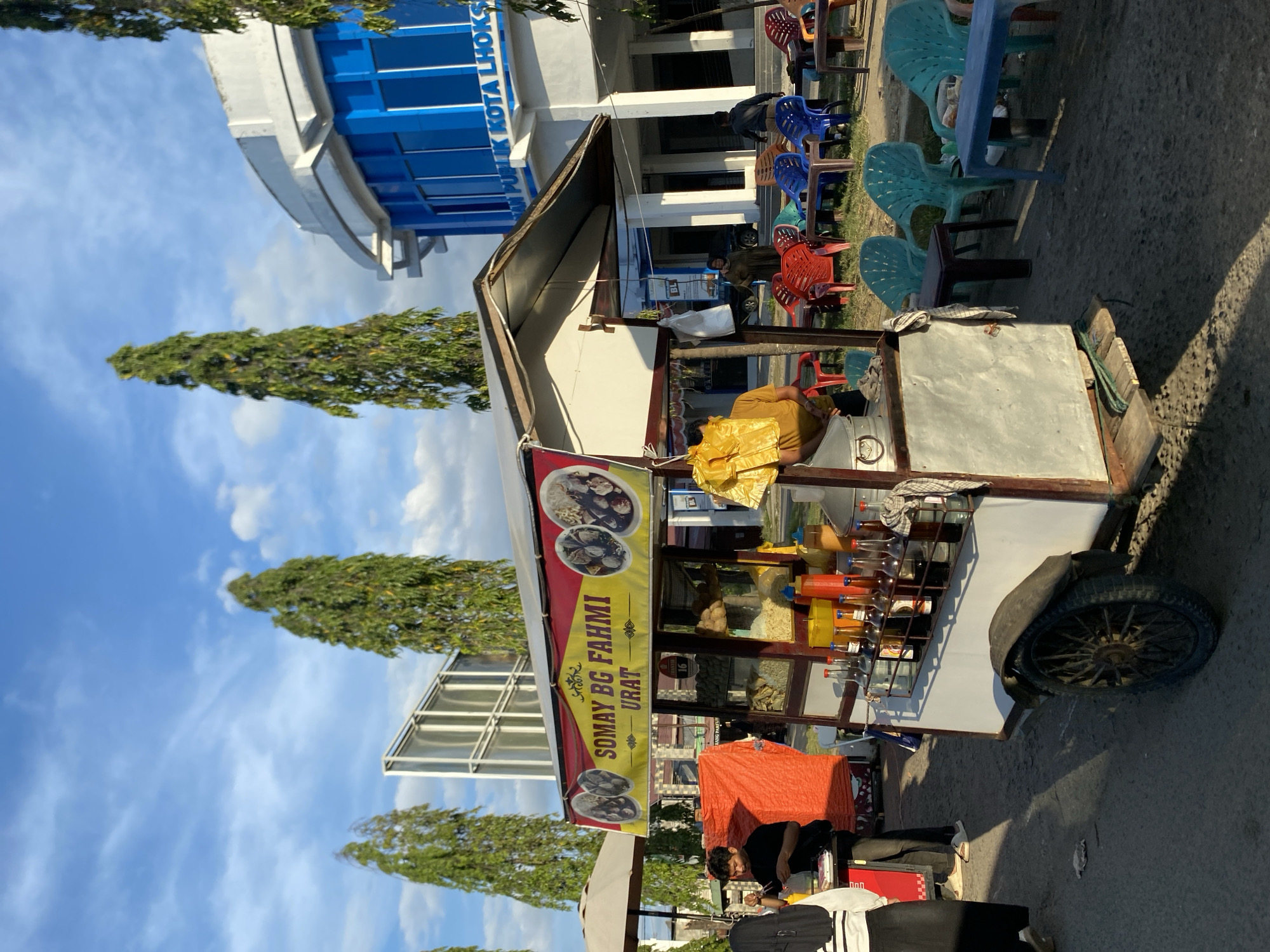 The image shows a street vendor's cart in Lhokseumawe, Indonesia. The cart is labeled "SOMAY BG FAHMI URAT" and displays pictures of the food being sold. A person is working at the cart, which is parked on a paved area with trees and buildings in the background. In the background, there is a building with blue and white features and several colorful chairs.