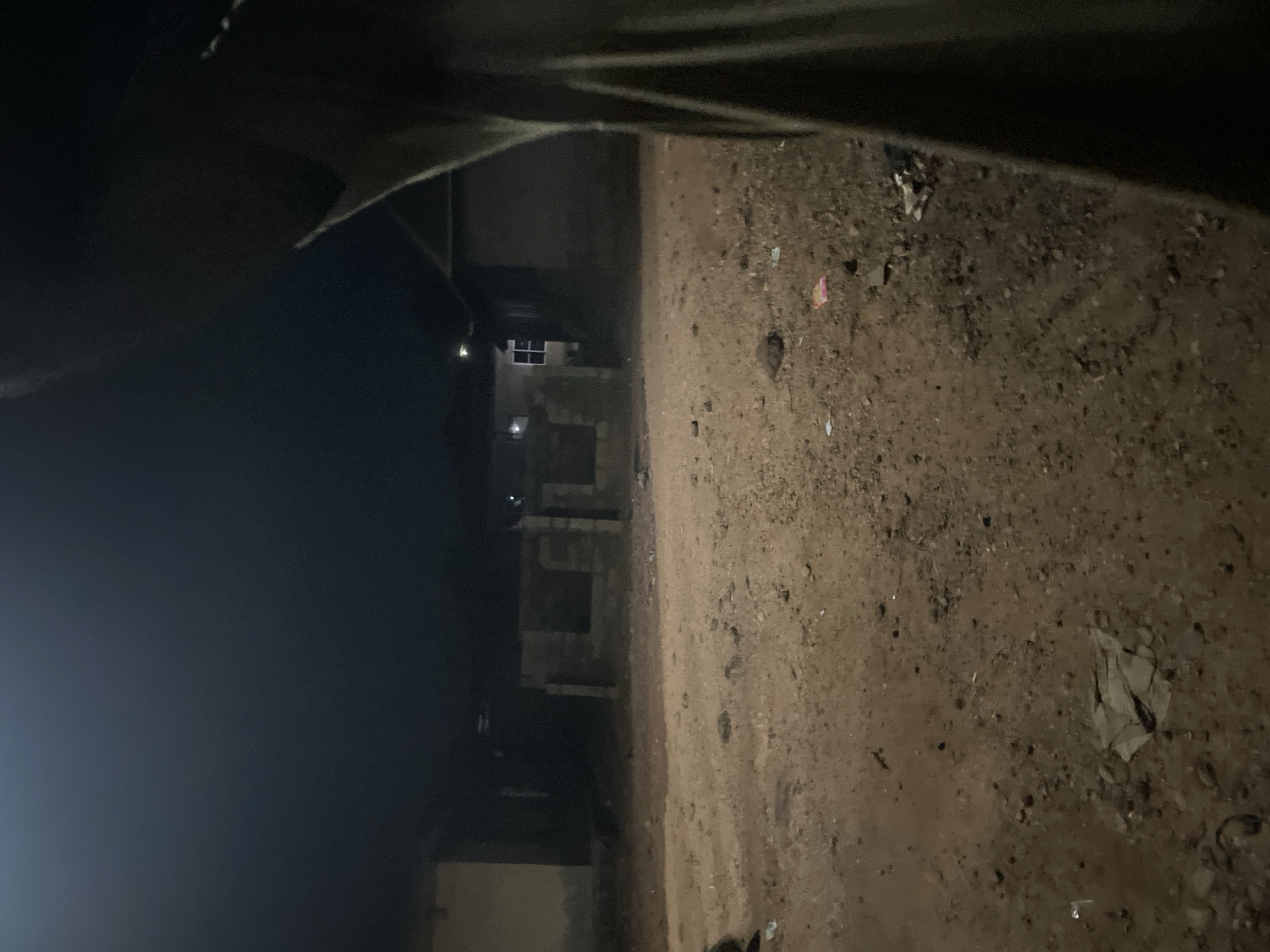 The image depicts an outdoor scene at night in Lokoja, Nigeria. The foreground consists of a dirt road with scattered pebbles and debris. In the mid-ground, there are partially constructed buildings or structures made of concrete blocks. One of these structures appears to have a window with a visible light inside. To the left, a more complete building with a light on is visible. The right side of the image is dominated by a dark, out-of-focus object, possibly a person's clothing or a curtain, partially obscuring the view. The overall atmosphere is dimly lit, characteristic of nighttime conditions.