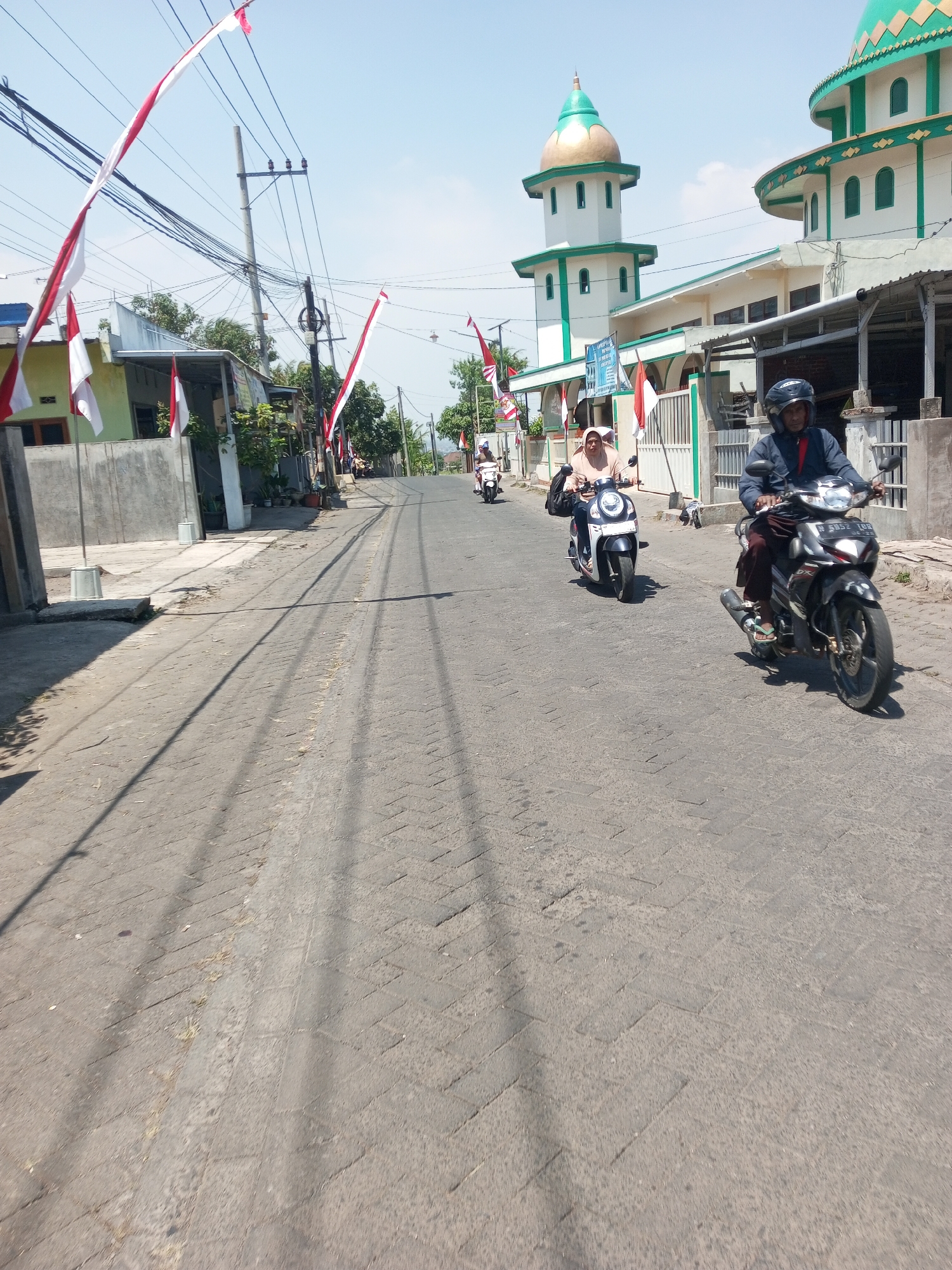 In Purwosari Klampok, Indonesia, a street scene unfolds under a bright sky. The road, paved with interlocking grey stones, is lined with buildings and utility poles adorned with Indonesian flags. In the foreground, two motorcycles are visible. The first, a black motorcycle with white accents, carries a person wearing a blue jacket and dark pants, with a black helmet. The second motorcycle, a white scooter, is ridden by a person in a light brown hijab and tan shirt. Further down the road, another motorcycle is seen with its rider. The background features buildings, including a mosque with a green dome and white walls, and houses. The atmosphere suggests a sunny day with a gentle breeze, as indicated by the fluttering flags.