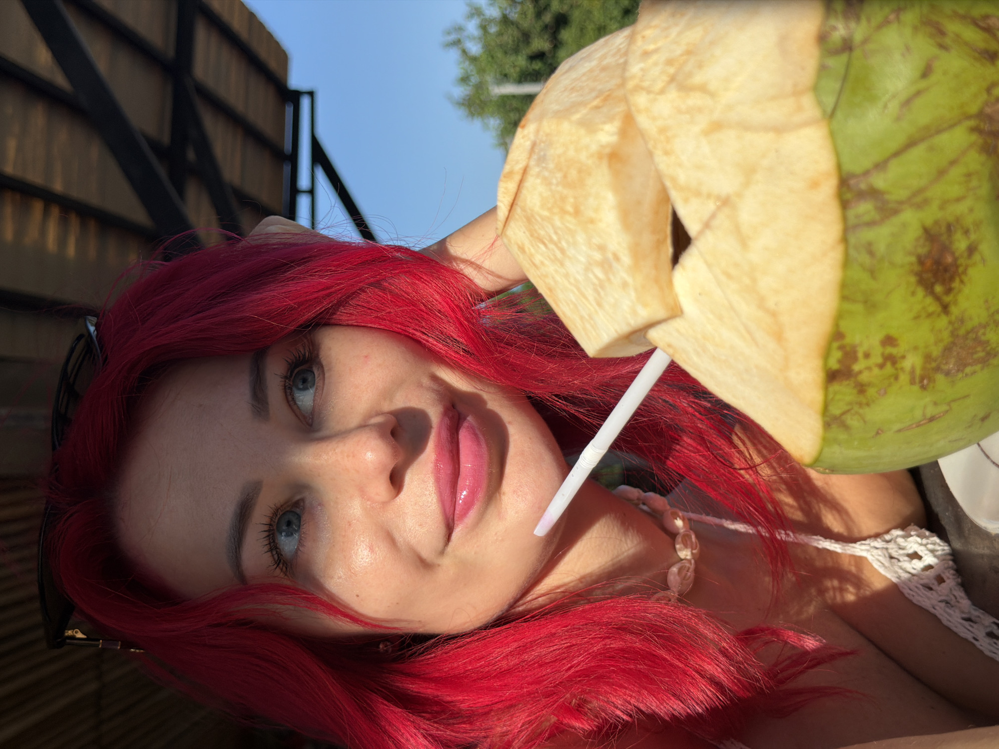 A close-up shot features a woman with vibrant, bright red hair and blue eyes looking to her right. She is wearing a white crocheted top and a shell necklace. Sunlight casts shadows across her face, highlighting her freckled skin and light pink lipstick. A white straw extends from her mouth, disappearing into a halved green coconut with a creamy white interior, which dominates the foreground of the image. The background suggests an outdoor setting with a thatched roof structure overhead and a bright blue sky visible. This scene is likely captured in Kuta, Indonesia, given the tropical ambiance suggested by the coconut and outdoor seating.