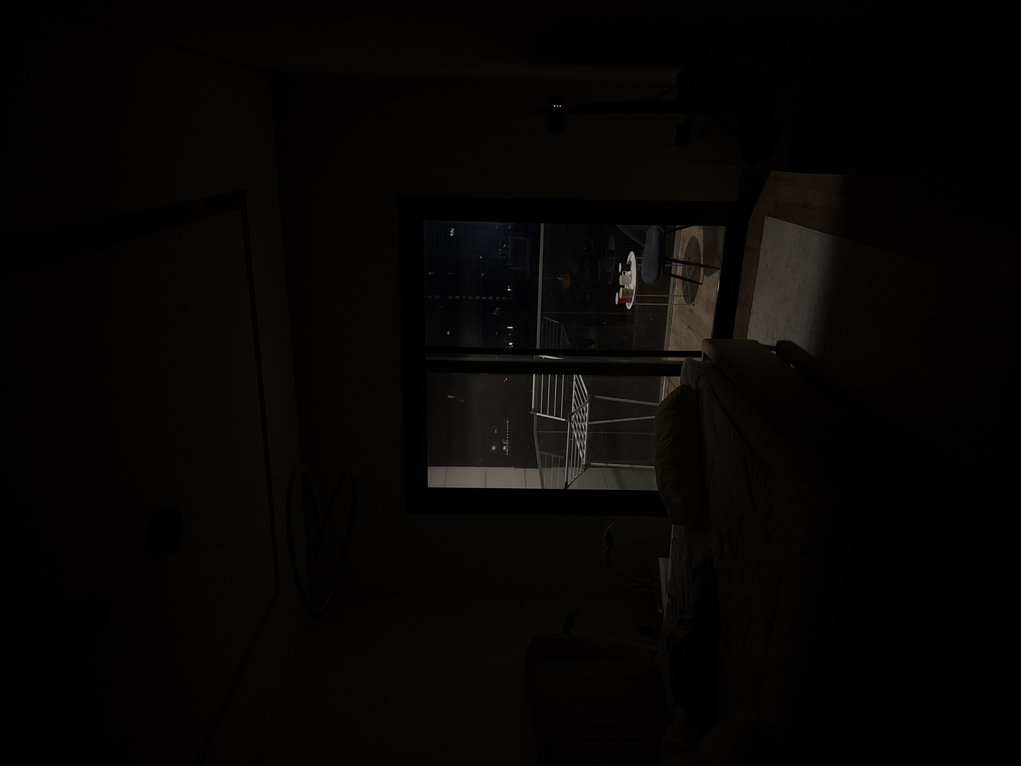 The image depicts a dark interior scene, likely a living room, with a large window overlooking a city at night. The city lights suggest an urban environment, and the prompt specifies the location as Dubai, United Arab Emirates. Inside the room, a white coffee table with a red object, possibly a cup or a candle, and a black chair are visible through the window. A bed or sofa is also present in the foreground, partially obscured by darkness. The overall mood is quiet and contemplative, possibly late at night. The limited lighting and the view of the illuminated city evoke a sense of urban isolation or peaceful observation.