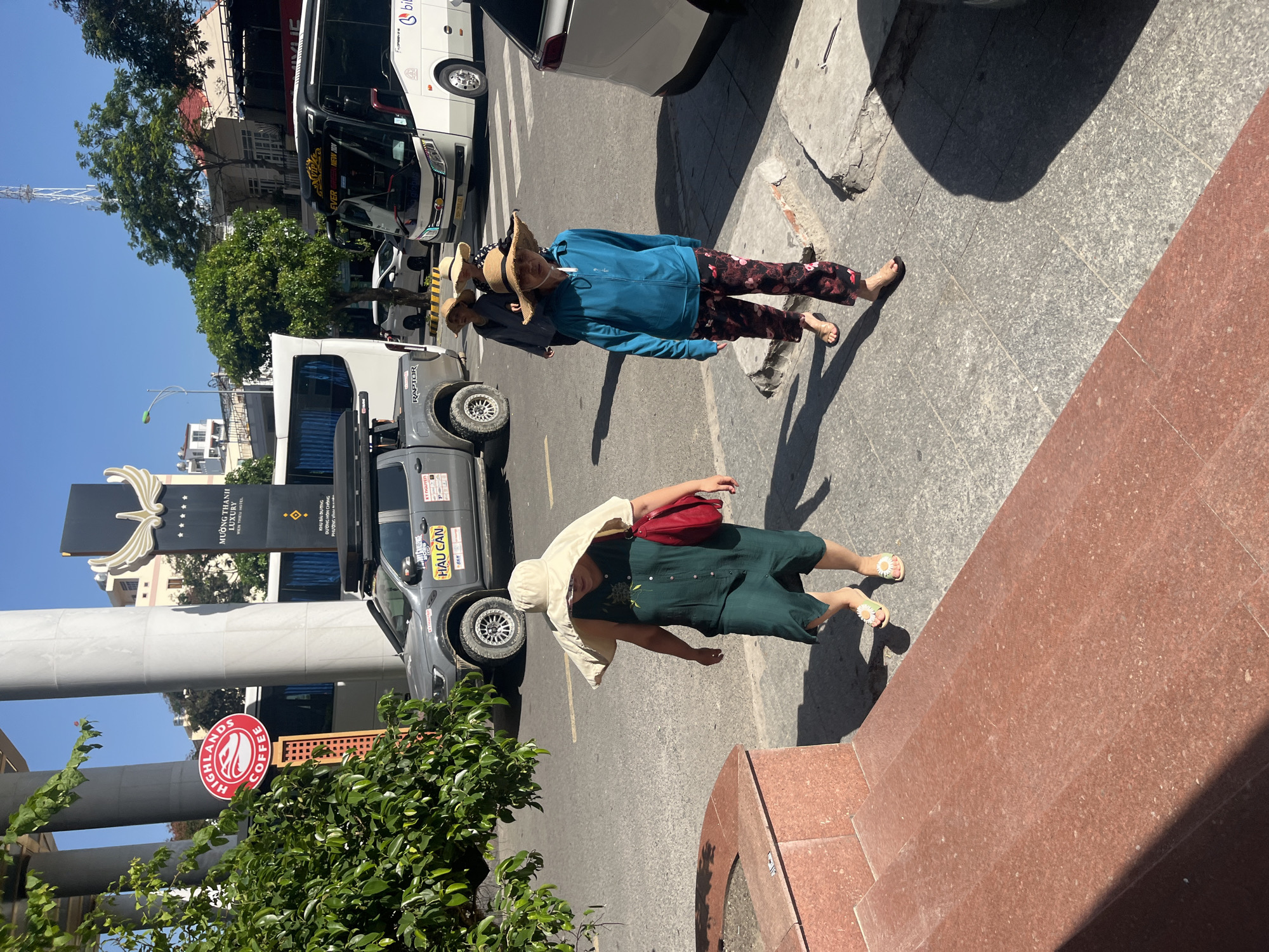 This image depicts a street scene in Bắc Nha Trang Ward, Nha Trang, Vietnam, identifiable by the prominent "MƯỜNG THANH LUXURY VIỄN TRIỀU HOTEL" and "HIGHLANDS COFFEE" signage. The scene is illuminated by bright daylight under a clear blue sky.
In the foreground, three women are walking from left to right on a grey speckled concrete sidewalk. The leftmost woman wears a wide-brimmed beige sun hat, a dark green sleeveless button-down dress with subtle embroidery, and light green flip-flops adorned with white flowers; she carries a red shoulder bag. The middle woman wears a straw hat with a checkered band and a dark top. The rightmost woman wears a straw hat with a black band, a long-sleeved blue zippered jacket, and dark floral pants (red and black pattern), with dark flip-flops.
To the left, a raised structure made of reddish-brown granite, possibly steps or a planter, partially frames the foreground. Green foliage partially obscures the "HIGHLANDS COFFEE" sign, a circular red sign with white text and a bird logo. Behind the foliage and coffee sign, large cylindrical columns of a building are visible, along with a tall, dark monument-style sign for the "MƯỜNG THANH LUXURY VIỄN TRIỀU HOTEL" featuring a golden winged emblem and five stars.
On the grey asphalt road, a grey Ford Ranger pickup truck with a rooftop tent and various stickers, including "HAU CAN" and "RAPTOR," is present. Behind it, a white passenger bus is visible. Further in the background, another white passenger bus, marked "EVEREST NEW 2024," faces the viewer, and a white SUV is partially visible on the far right. A white cellular tower or antenna structure stands against the sky in the distant background. Shadows from people and objects are cast on the ground, indicating strong sunlight.