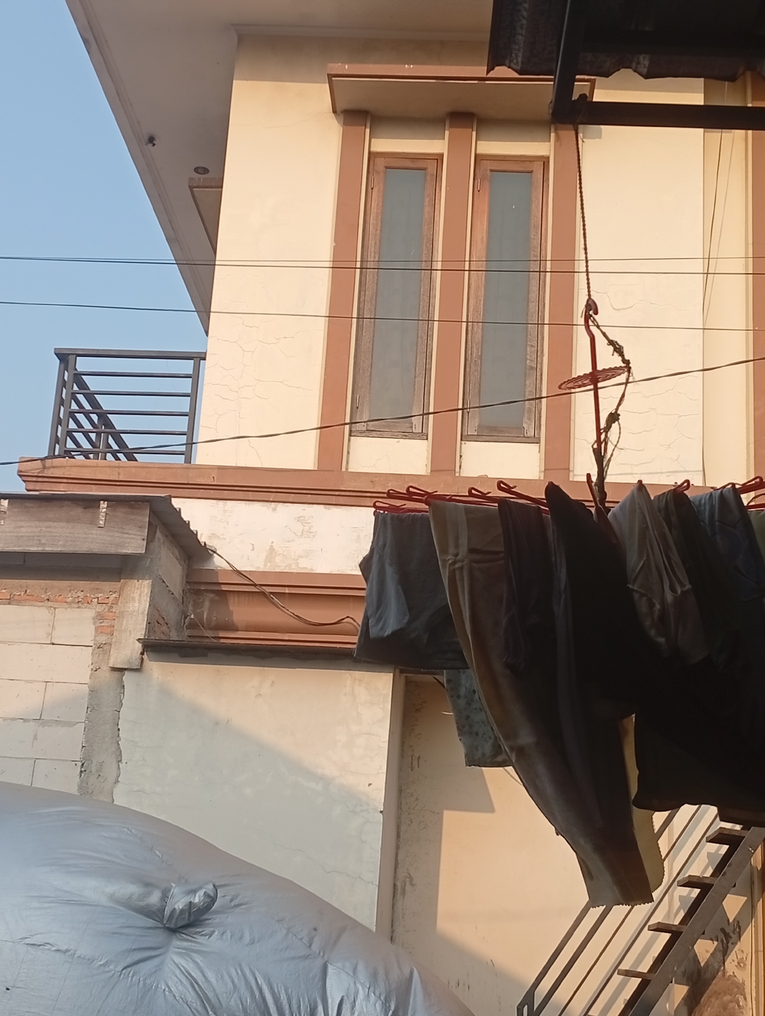 The image depicts an outdoor scene in Bekasi, Indonesia, likely on a residential property. In the foreground, laundry is hung on a clothesline, with several garments including pants and shirts visible. The laundry is attached to a red circular drying rack mechanism suspended by a rope from an overhang. Behind the laundry, a portion of a two-story building is visible. The building has a cream-colored facade with brown trim around two tall, narrow windows. A balcony railing made of dark metal bars is attached to the side of the upper floor. To the left of the building, a wall constructed of light-colored cinder blocks is partially visible. In the lower left corner of the image, a large, silver-colored object, possibly a tarped item or a large bag, is present. A metal staircase leading upwards is visible on the right side of the image. The sky is a clear light blue, suggesting it is daytime. The overall scene suggests a domestic setting where laundry is being dried.