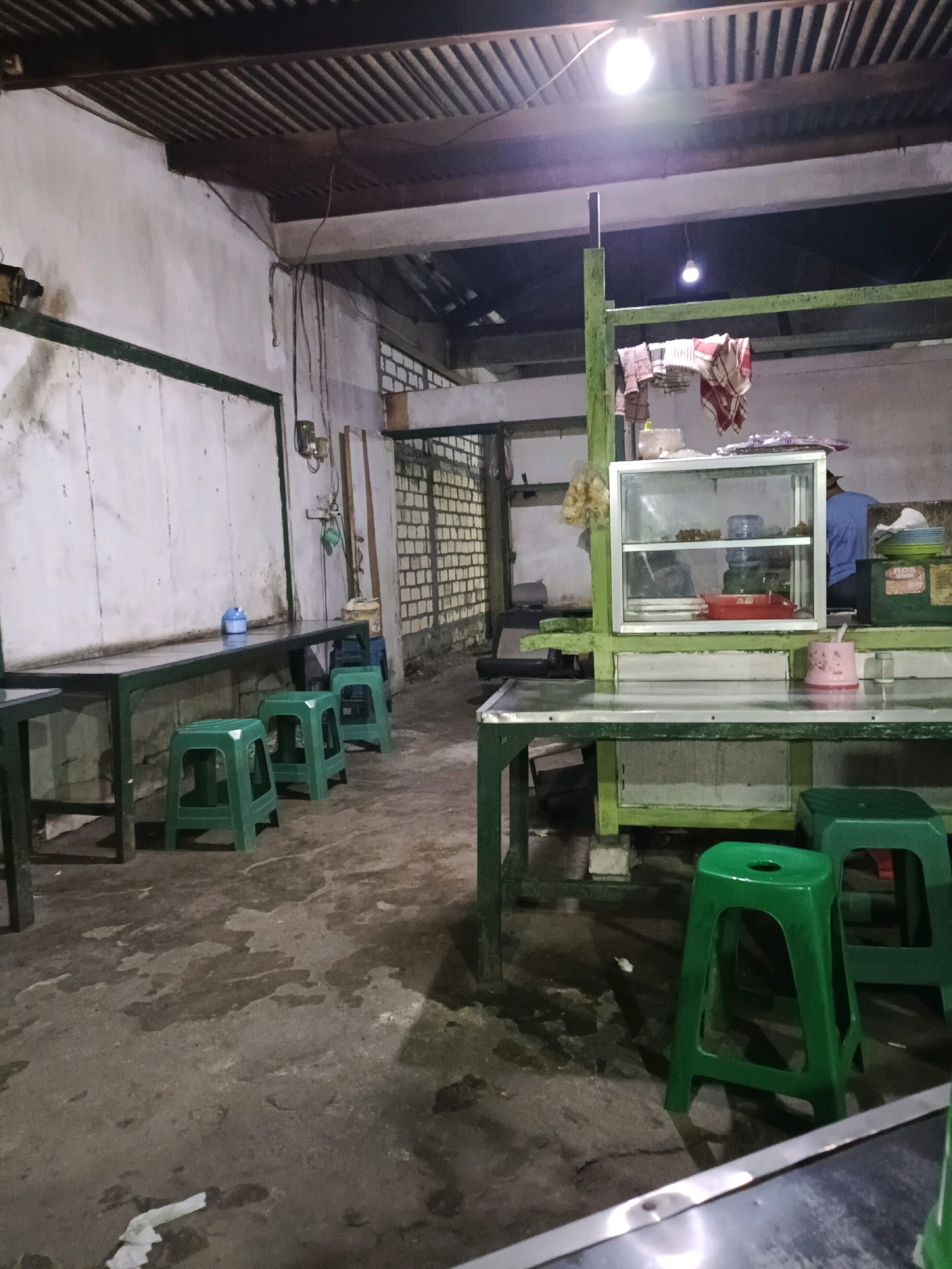 The image depicts the interior of a roadside food stall in Sukomulyo, Indonesia, at night. The stall features several green plastic stools arranged around metal tables. A glass display case is set up on one of the tables, containing food items. A person wearing a blue shirt is partially visible in the background. The environment appears somewhat rustic, with a corrugated metal roof and concrete flooring. The lighting is dim, with a bright overhead light illuminating the scene. Laundry is hanging on a rack in the background.