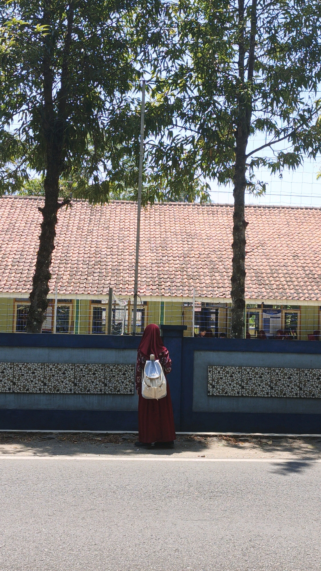 A person wearing a maroon hijab and a maroon patterned dress, with a silver backpack, stands on the sidewalk facing away from the camera. Behind the person is a blue fence with a decorative stone pattern. Beyond the fence is a building with a red tile roof and green and yellow walls, featuring several windows. Two trees with green foliage are on either side of the building, obscuring parts of the sky. The scene is located in Cibenda, Indonesia, under a clear blue sky. A paved road is in the foreground.
