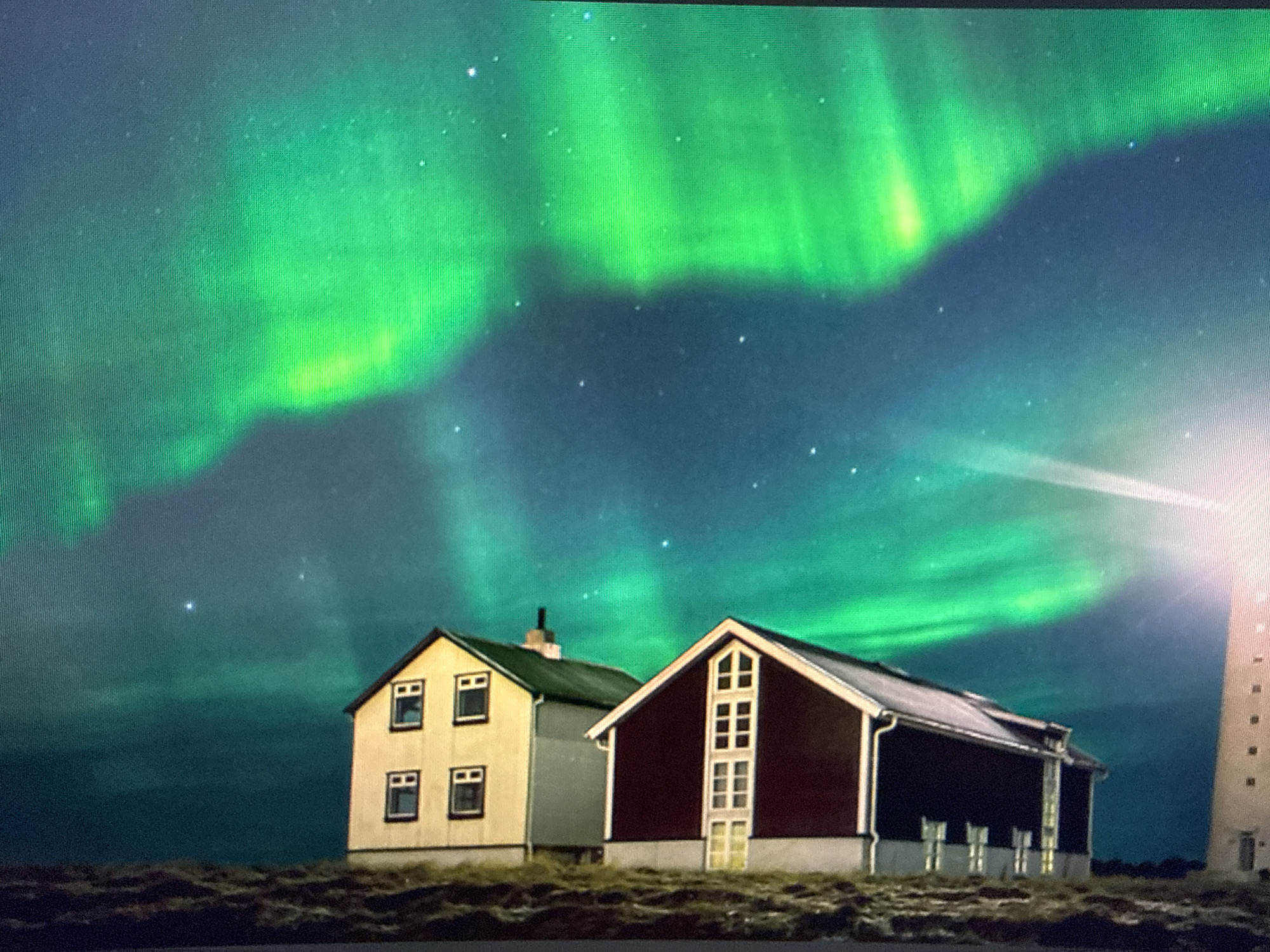 The image depicts a night scene with the aurora borealis (Northern Lights) visible in the sky. Two buildings are in the foreground: a light-colored building with four visible windows, and a darker red building with white trim and multiple windows on one side. A tall, white lighthouse is partially visible to the right, emitting a bright beam of light. The ground appears to be grassy or covered in low vegetation. There are visible stars in the sky behind the aurora. The location is not in Sofia, Bulgaria, but rather in a location where the aurora borealis is visible, such as Iceland or Norway.