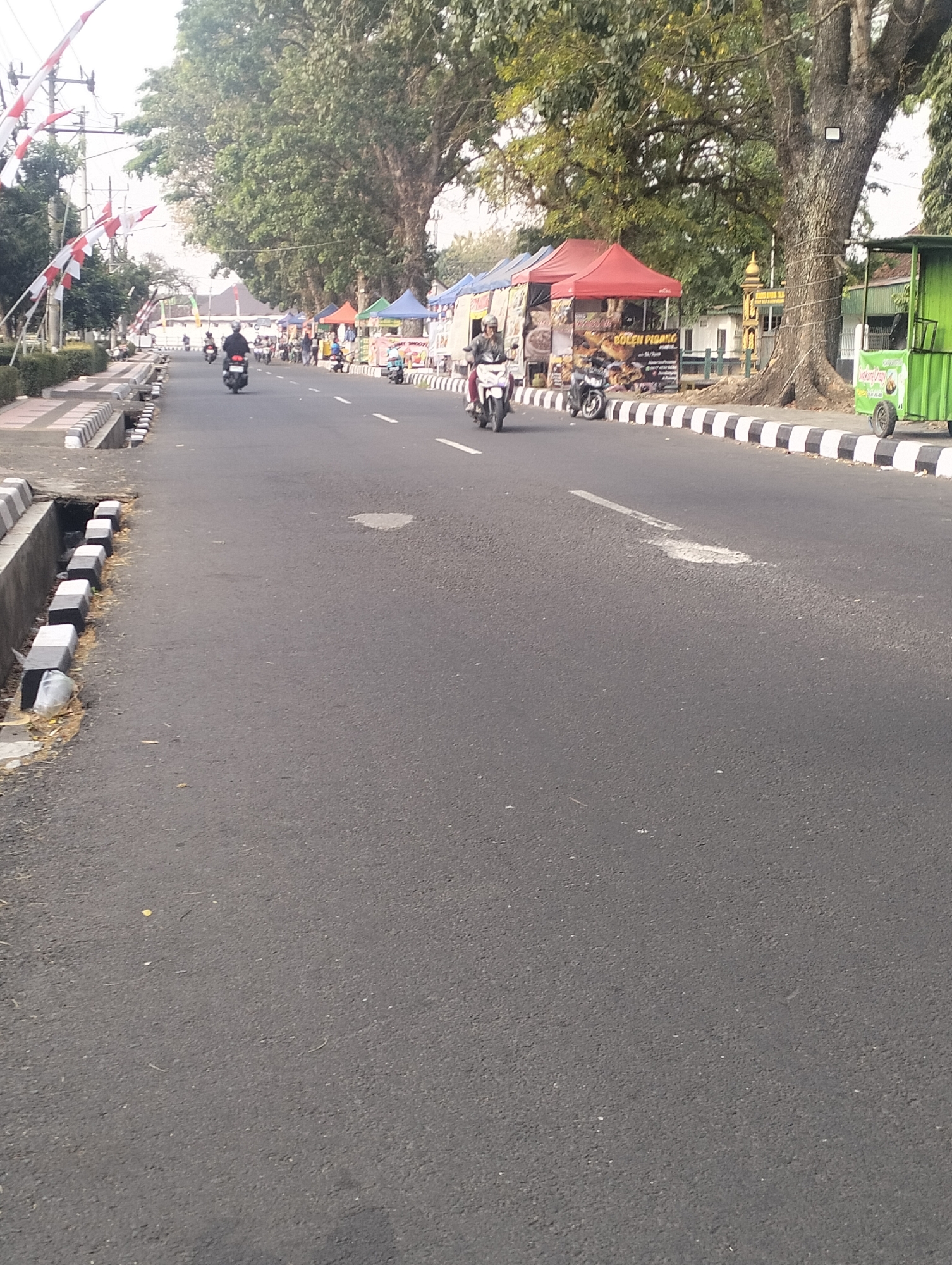 The image captures a street scene in Magelang, Indonesia, characterized by a gray asphalt road. On the left side of the road, a concrete sidewalk with black and white striped curbing lines a drainage ditch. Several red and white Indonesian flags are displayed on poles along the sidewalk. Further down the road, on the right side, a row of temporary stalls with colorful awnings, predominantly red and blue, are set up. These stalls appear to be selling food or goods. A large, mature tree with dense foliage stands prominently behind these stalls. A bright green mobile cart with wheels is parked near the stalls. Several motorcycles are present, with some riders actively on the road. One motorcycle in the foreground on the right shows a rider. Another rider on a motorcycle is visible in the middle ground, moving away from the camera. The overall environment suggests a lively street with commercial activity, likely during the daytime, given the natural lighting.