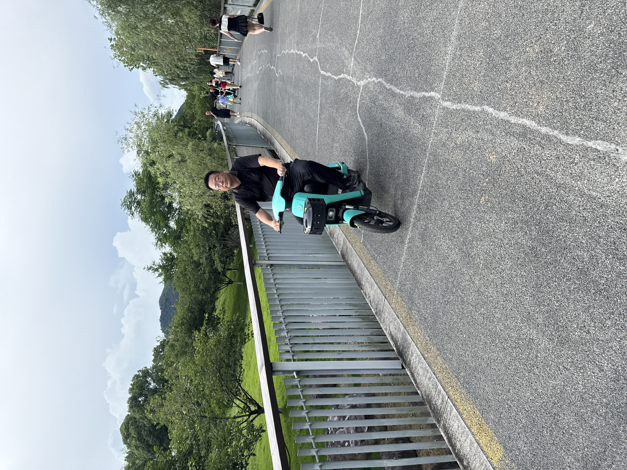 A man in a black polo shirt and black pants rides a teal electric scooter forward on a paved pedestrian path in Lin'an District, China. He is wearing glasses and looking ahead. The path is bordered on the left by a metal railing and a grassy slope with trees. To the right of the path, other people are walking, including a child in a blue shirt and yellow shorts. Trees and foliage line the sides of the path, with a cloudy sky overhead. The pavement has a light gray speckled texture with white lines marking the path.