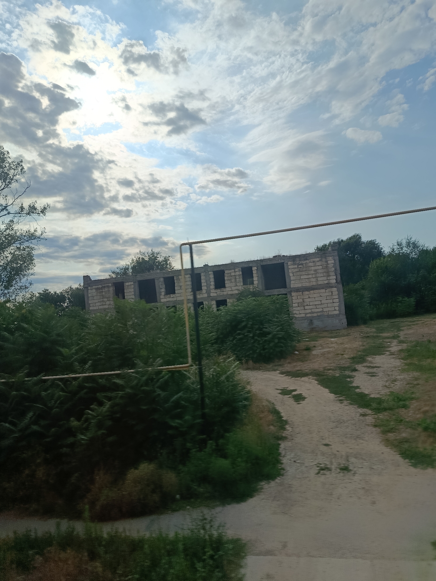 The image shows an unfinished concrete building with multiple empty window openings, surrounded by lush green vegetation. A dirt path leads towards the building. In the foreground, there is more dense greenery, and a thin yellow pipe runs horizontally across the scene in front of the building. The sky above is partly cloudy with the sun shining through. The location is identified as Congaz, Moldova. There are no people visible in the image. The scene appears to depict an abandoned or under-construction structure.