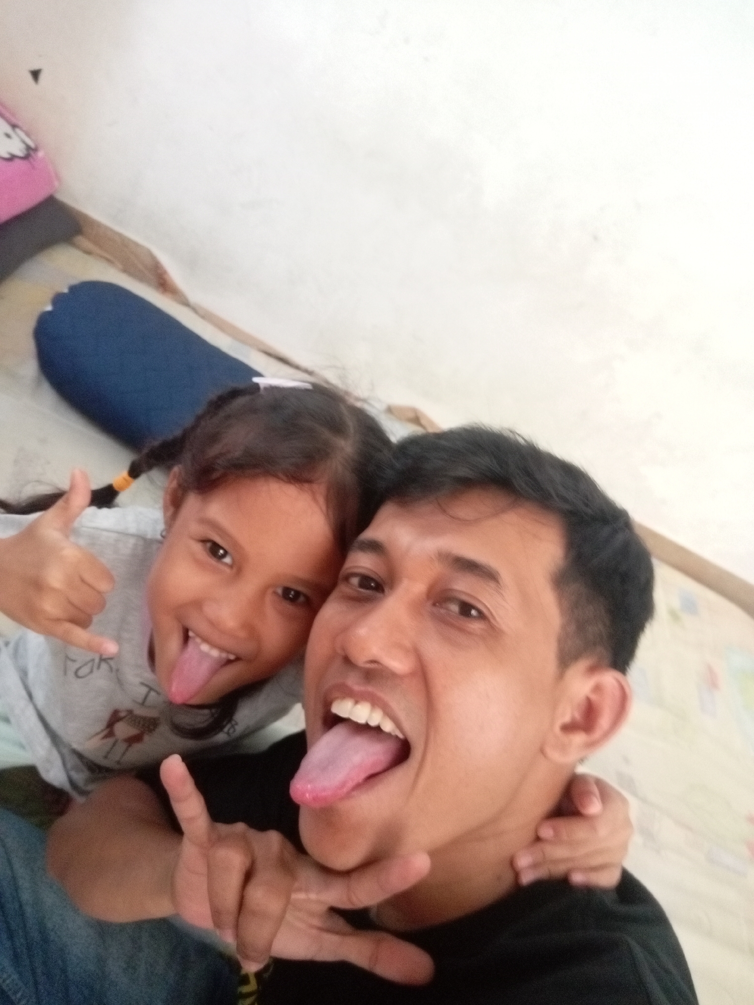 A young girl and a man are taking a selfie. Both are sticking out their tongues and making playful hand gestures. The girl has her thumb up and her pinky extended, while the man is making a "rock on" sign with his hand. The girl has her hair in pigtails with orange ties. She is wearing a gray shirt with some text and a drawing. The man is wearing a black shirt. They appear to be indoors, possibly in a home, with a white wall in the background and some colorful items on the floor. The lighting suggests it could be daytime. The overall mood is joyful and lighthearted.