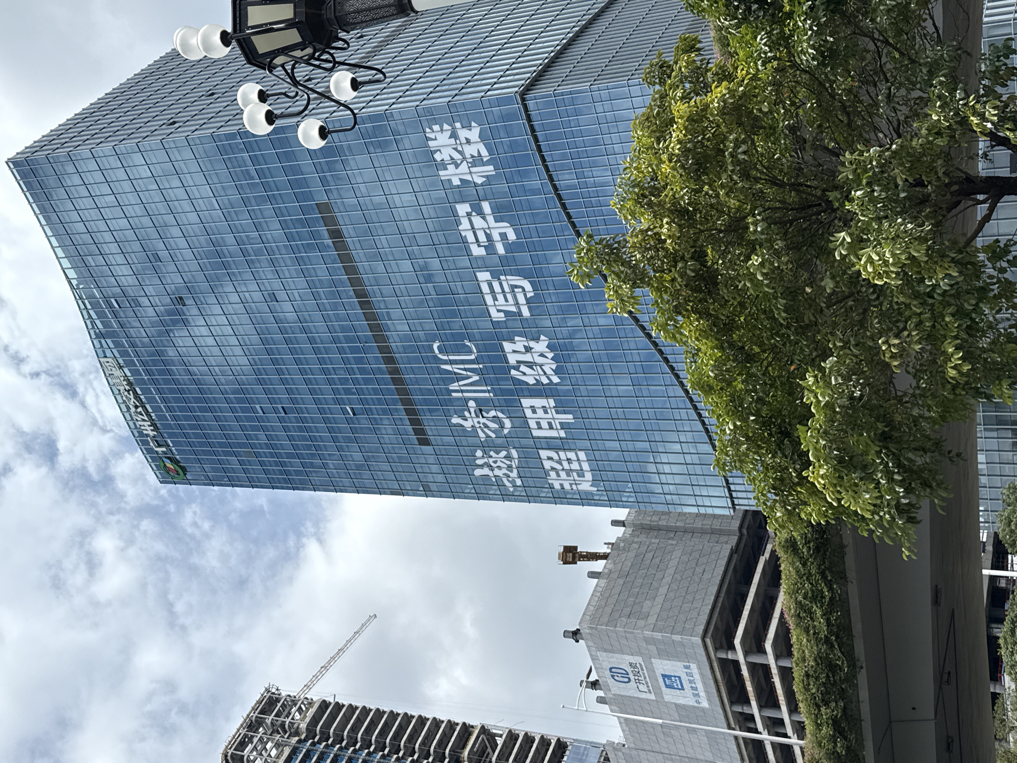 A high-angle, medium shot captures a modern cityscape in Guangzhou, China, focusing on the Huangpu District. The primary subject is a tall, glass-facaded skyscraper with white lettering across its surface. The lettering appears to be a slogan or advertisement. To the left of this skyscraper, another building under construction is visible, with its structural framework exposed and a crane extending upwards. In the foreground, lush green trees obscure the lower portions of the buildings and an elevated roadway. The sky is overcast with clouds, suggesting a temperate climate. The scene is devoid of any human presence.