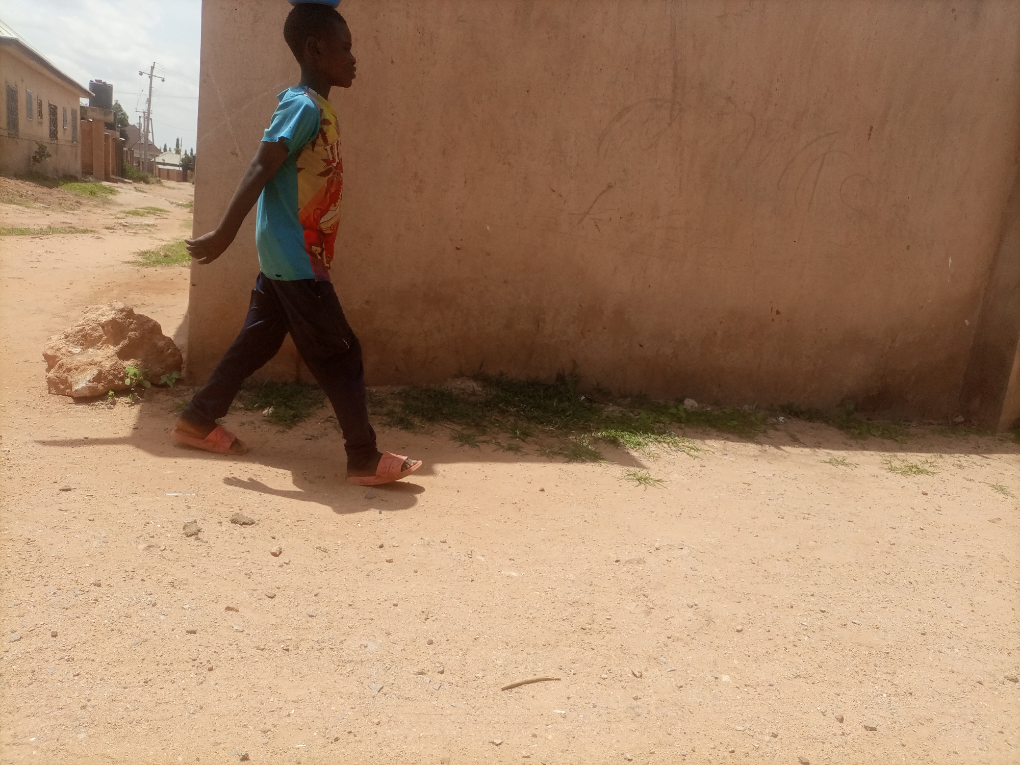 A young boy, carrying a blue object on his head, walks on a dirt road in front of a light brown wall. He is wearing a blue and red t-shirt and dark pants. His feet are bare except for pink sandals. The road is sandy with some small rocks and patches of grass. In the background, there are buildings and a utility pole. The overall environment suggests a rural or semi-urban setting in Tumfure, Nigeria. The lighting indicates it is daytime, with shadows cast on the ground. The wall appears to be made of adobe or a similar material and has some faint markings on its surface.