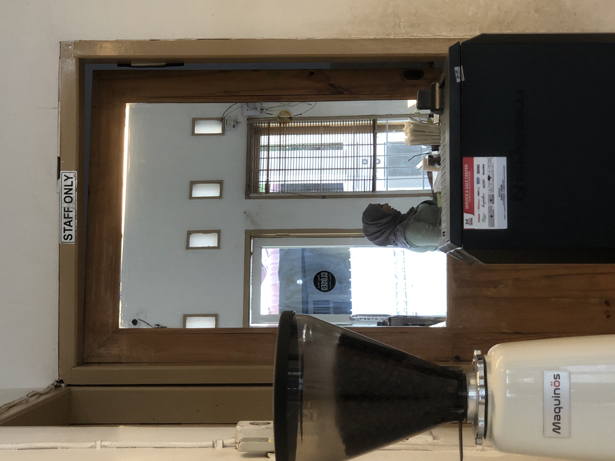 The image captures a scene in a building, seemingly a cafe or restaurant, in Lhokseumawe, Indonesia. A woman wearing a hijab is visible in the background, behind a counter. The environment includes a window with blinds and several small rectangular openings in the wall. A black machine with a sticker displaying text and logos related to service and sale center is positioned on the right. In the foreground, part of a Maquinos coffee grinder is visible. A "STAFF ONLY" sign is affixed to the side of a doorway.