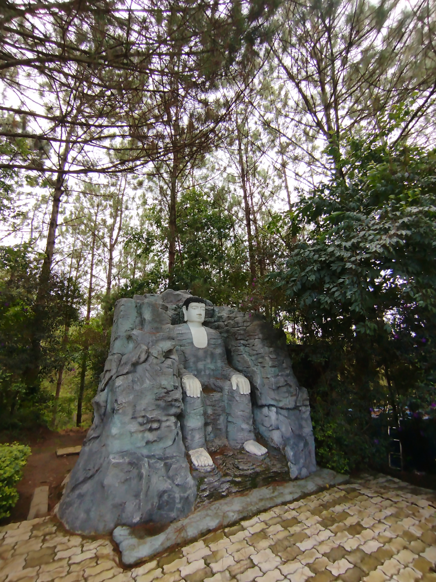 The image captures a statue of a figure seated in a meditative pose, possibly Buddha, integrated into a rocky structure. The statue has a white head and white hands and feet. The rest of the statue is made of gray material. The structure is placed outdoors amidst dense foliage, including tall pine trees. The ground is paved with interlocking bricks. The scene is located in the city of Lam Vien - Da Lat, Vietnam.