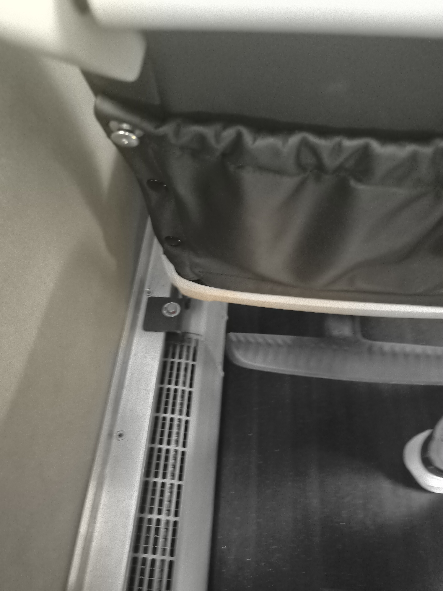 This image shows a close-up of a section of a treadmill. The left side features a vertical ventilation grill set into a silver-colored metal frame. Above this grill, a black cloth panel is attached, possibly a safety barrier. The cloth is gathered at the top. A white or light-colored bar or edge runs horizontally, possibly a component of the treadmill's frame or control panel. The treadmill belt is visible as a dark, flat surface. Part of a shoe is visible on the right side of the image. The background includes what appears to be a light gray wall.
There is no indication of L'Horme, France in this image.