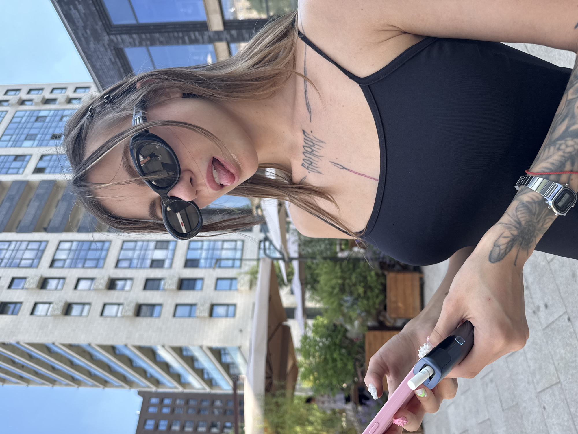 A young woman with long, light brown hair, wearing large dark sunglasses and a black tank top, sticks her tongue out playfully at the camera. She has tattoos on her chest and left arm, and wears a digital wristwatch on her right wrist. Her hair is partially pulled back with metallic hair clips. She is holding a pink phone case and a dark blue and light blue electronic device in her hands. The background shows a modern, light-colored building with many windows, suggesting an urban setting. Green trees and outdoor seating areas are also visible, hinting at a public space like a cafe or park. The lighting suggests it is daytime, likely sunny. The overall mood appears casual and lighthearted. No visible text is present. The location is Moscow, Russia.