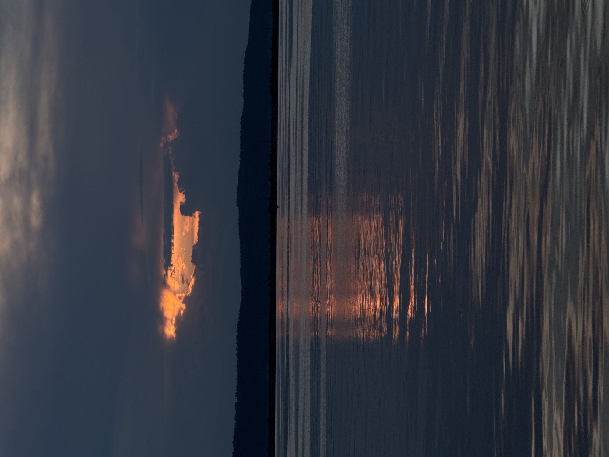 A wide shot captures a serene body of water, likely the Kama River, near Chaikovsky, Russia, under a dramatic twilight sky. The water's surface is relatively calm, with subtle ripples reflecting the fading light. A dark, silhouetted treeline forms the distant horizon, interrupted by what appear to be faint vertical structures, possibly utility poles or pylons.
The sky is a canvas of dark, brooding clouds, but a break in the overcast reveals a vibrant patch of orange and yellow light, suggesting the last rays of the sun piercing through. This intense light is mirrored on the water's surface, creating a striking streak of warm color against the cool, dark tones of the river. The overall impression is one of peaceful solitude and the raw beauty of nature at dusk. There are no people or specific man-made objects in the immediate foreground or visible on the water's surface, emphasizing the natural landscape.