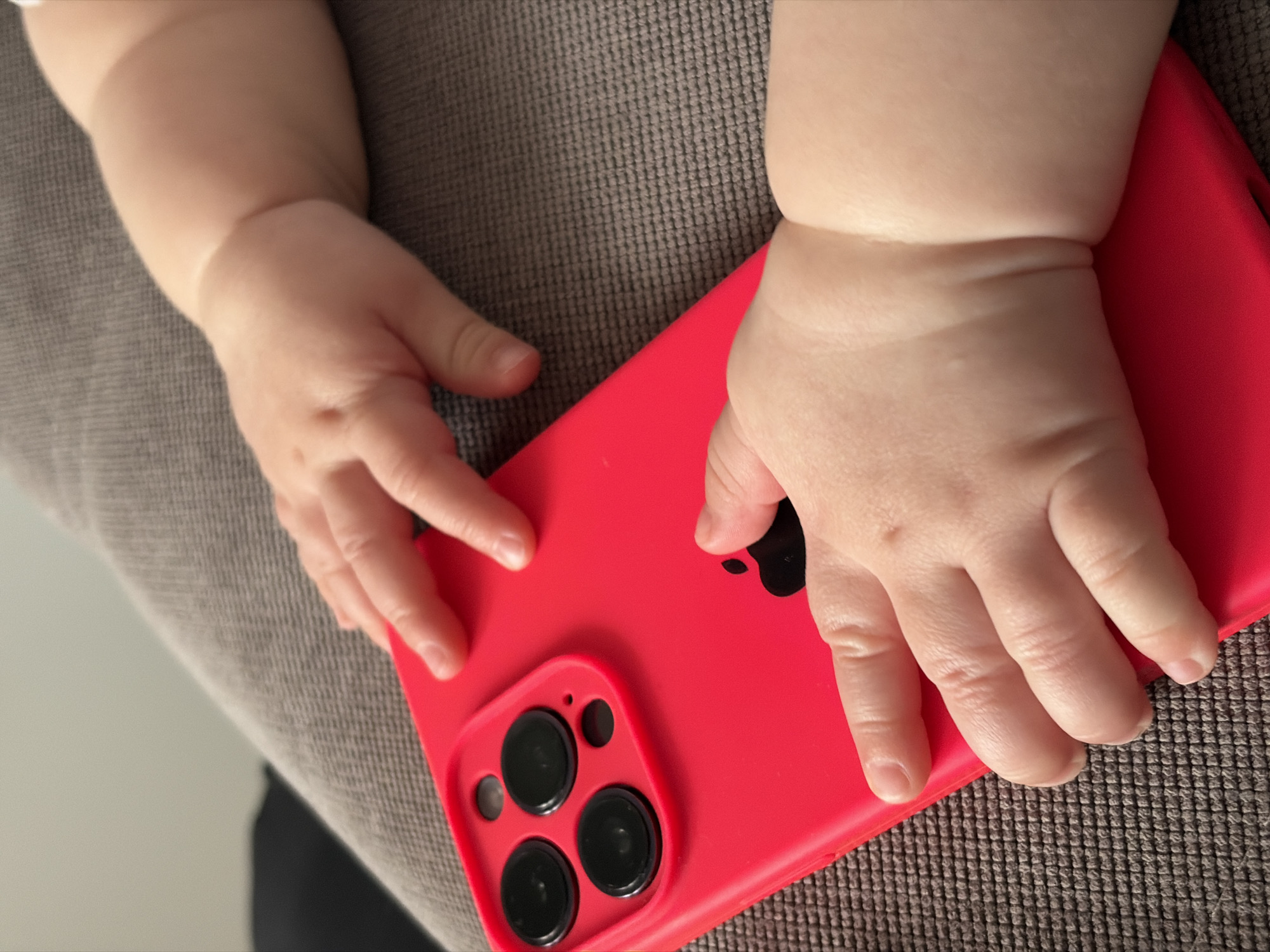 The image shows a red phone, likely an iPhone, lying on a grey fabric surface. A baby's hands are resting on the phone. The hands appear chubby and soft. The camera of the phone is visible with three lens and one small circle probably for the flash. The background is a textured fabric, possibly a sofa or blanket. The image doesn't contain any evidence to confirm that this image was taken in Krasnodar, Russia. Therefore, this information cannot be confirmed.
