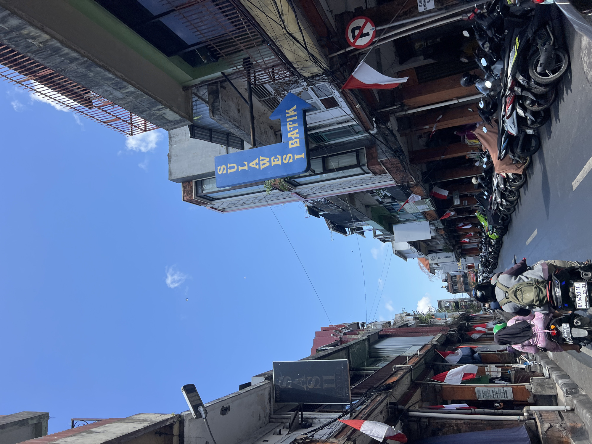 In Denpasar, Indonesia, a street scene unfolds under a clear blue sky with a few wispy clouds. The image captures a narrow street lined with multi-story buildings, likely shops or businesses, displaying an assortment of signage and decorations. Indonesian flags, red and white, are prominently featured, hung from poles and storefronts, suggesting a national celebration or patriotic display.
On the left side of the street, a black rectangular sign with white lettering reads "SAS II TEXTILE," indicating a business that sells textiles. A street lamp is affixed to the building above this sign. Further down, another building displays a blue sign with white lettering that says "SULAWESI BATIK" with a blue arrow pointing to the right, directing towards a batik shop. A "no parking" sign with a red circle and a diagonal line over a red 'P' is visible on the right side of the street.
The street itself is paved and appears to be a busy thoroughfare, with numerous motorcycles parked along the right side of the road. Two individuals are riding a motorcycle in the foreground, the passenger wearing a pink garment and a black headscarf, and the driver wearing a backpack. They are moving away from the viewer down the street. The buildings exhibit various architectural styles, some appearing older and weathered with exposed concrete and metalwork, while others have more modern facades with glass windows. Power lines and cables are strung across the street between the buildings, creating a complex network. The overall impression is of a vibrant commercial street in a Southeast Asian city, with a mix of traditional and contemporary elements.