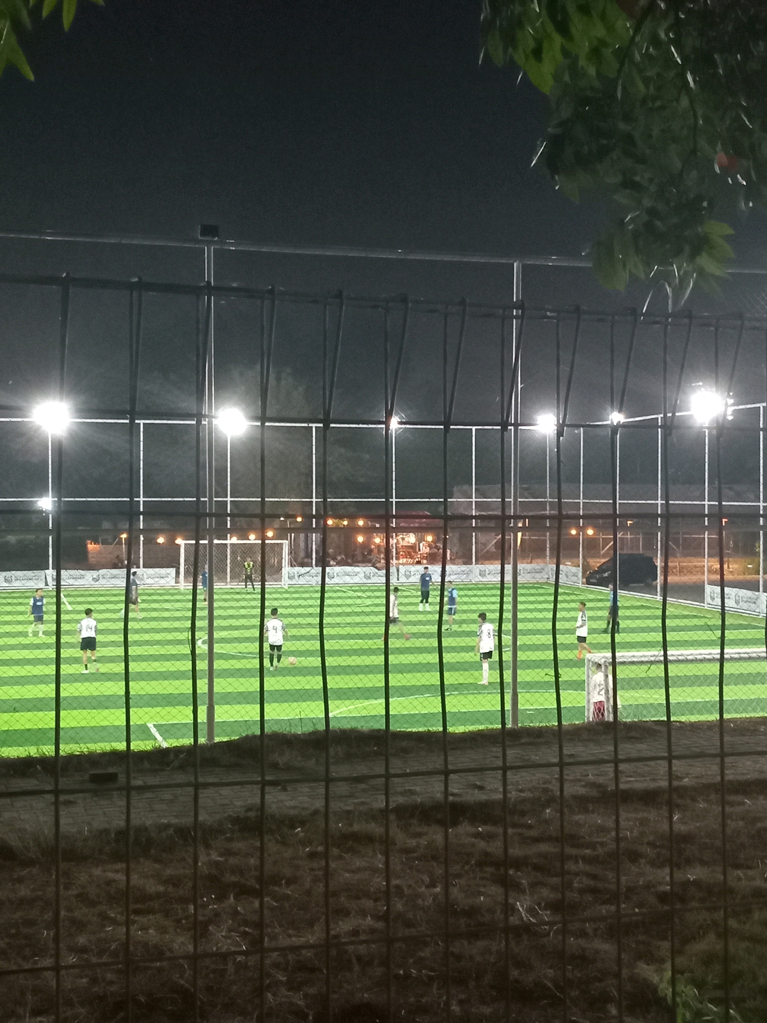This is a night time photograph taken from behind a fence, looking onto a brightly lit artificial turf soccer field where a game is in progress. Several individuals, presumably players, are scattered across the field in athletic attire, some in white and black uniforms, others in blue. One individual, likely a goalkeeper, is positioned near a soccer goal. The field is marked with white lines and illuminated by overhead floodlights. In the background, beyond the field, there are buildings with lights, possibly a viewing area or amenities, and a dark colored vehicle. The foreground shows the fence and dry grass or dirt. The presence of a soccer field with lights suggests it is a sports facility. The location is identified as Karangturi, Indonesia.