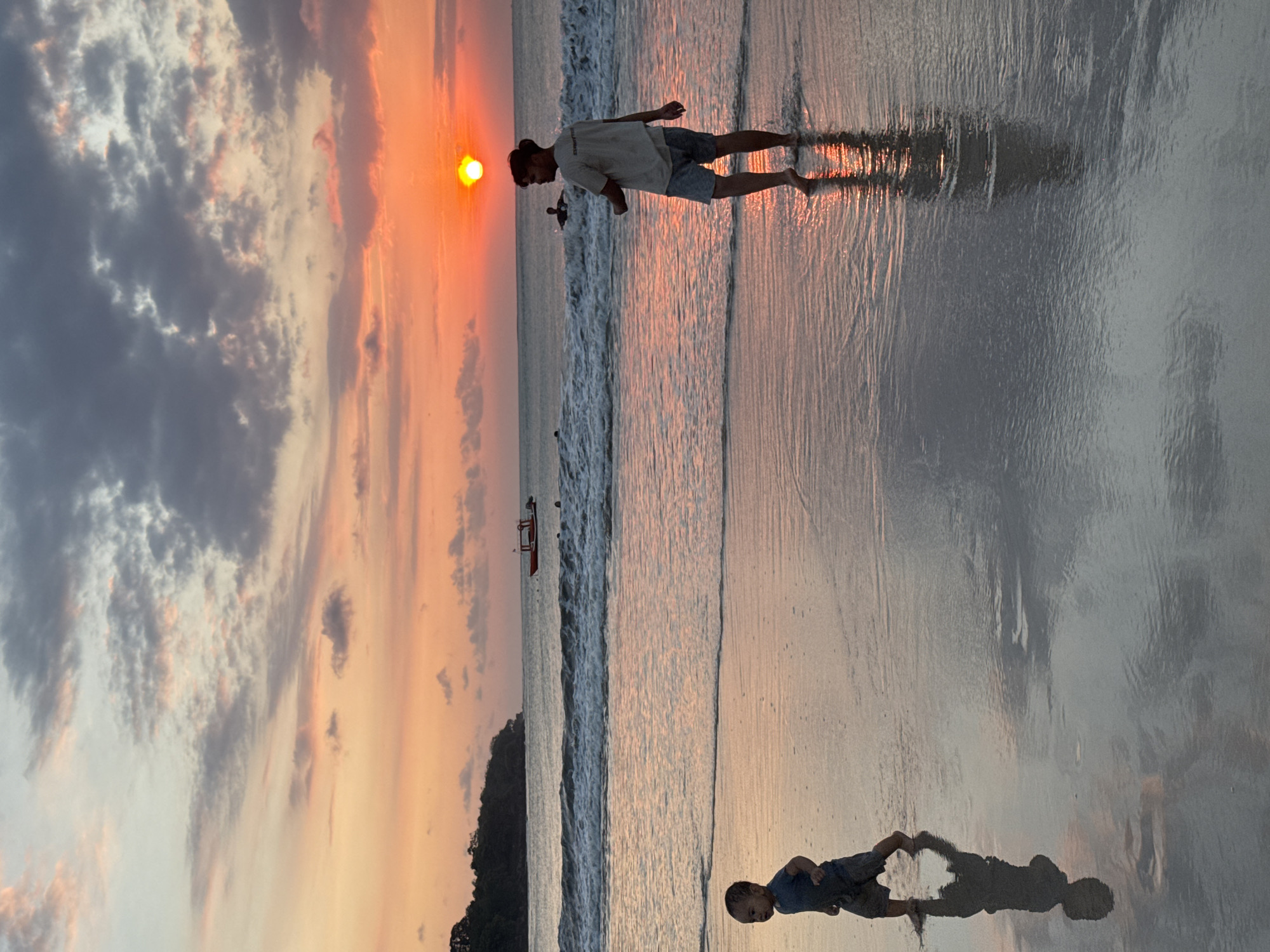 The image depicts a tranquil sunset scene on a beach, characteristic of a coastal location like Kuta Selatan, Indonesia. The sun, a vibrant orange-yellow sphere, is positioned low on the horizon to the left, bathing the entire setting in a warm, golden light.
In the foreground, a young man stands barefoot on the wet, reflective sand, wearing a light-colored t-shirt and shorts. He is near the water's edge, where gentle waves roll in. His clear reflection is cast upon the glistening sand behind him. Further to the right in the foreground, a young child is also present on the wet sand, facing the ocean, with their distinct shadow and reflection visible. In the distance, another person is partially silhouetted in the water, and a small, traditional red and white boat floats peacefully further out.
The sky is a captivating blend of sunset colors, with soft oranges, pinks, and yellows merging with grays and blues of scattered clouds. A dark, hilly landmass, possibly a distant island or part of the coastline, is visible between the ocean and the sky on the right side. The wet sand acts as a natural mirror, beautifully reflecting the radiant sky, the breaking waves, and the figures on the beach. The scene evokes a sense of peace and natural beauty at dusk. There is no visible text in the image.