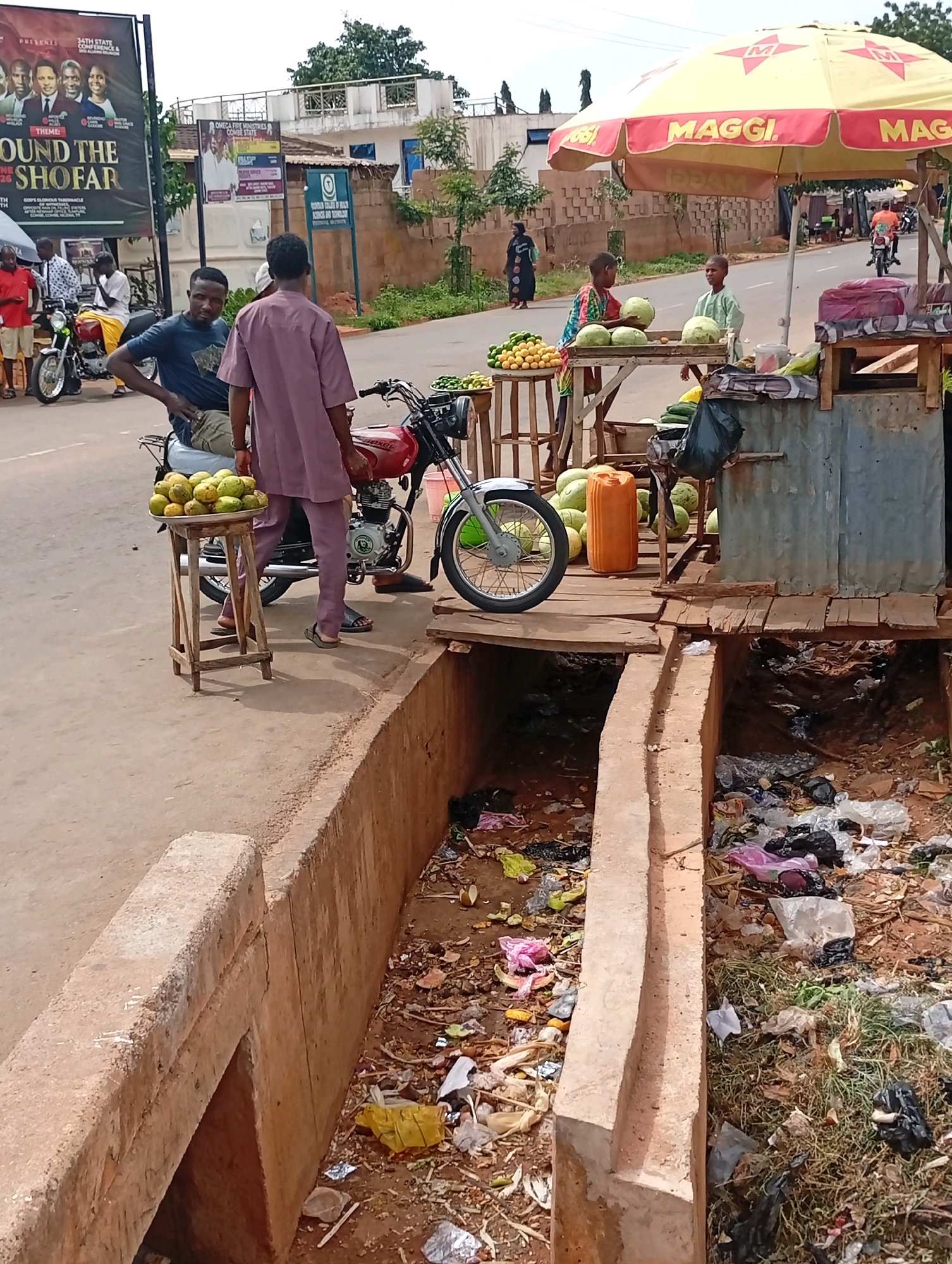 The image depicts a street scene in Tempure, Nigeria, with a focus on a roadside fruit stand and its surrounding environment. In the foreground, a concrete drainage ditch filled with various types of litter, including plastic bags, wrappers, and organic waste, runs along the side of the road.
Adjacent to the ditch is a paved road where several individuals are present. Two men are standing near a red motorcycle. One man, wearing a blue t-shirt and dark pants, is seated on the motorcycle and looking towards the viewer. The other man, dressed in a purple outfit, stands beside him, also near the motorcycle. A tray of what appear to be mangoes sits on a wooden stool in front of the motorcycle.
Further down the road, under a large yellow umbrella with "MAGGI" branding, a small fruit stand is set up. Two young boys are interacting at the stand, which displays watermelons and other fruits. A tall orange plastic container is also visible. In the background, more people and vehicles, including motorcycles, can be seen. Buildings and vegetation form the backdrop of the scene, with a large banner visible on the left side of the image. The overall impression is a bustling but somewhat unkempt urban environment.