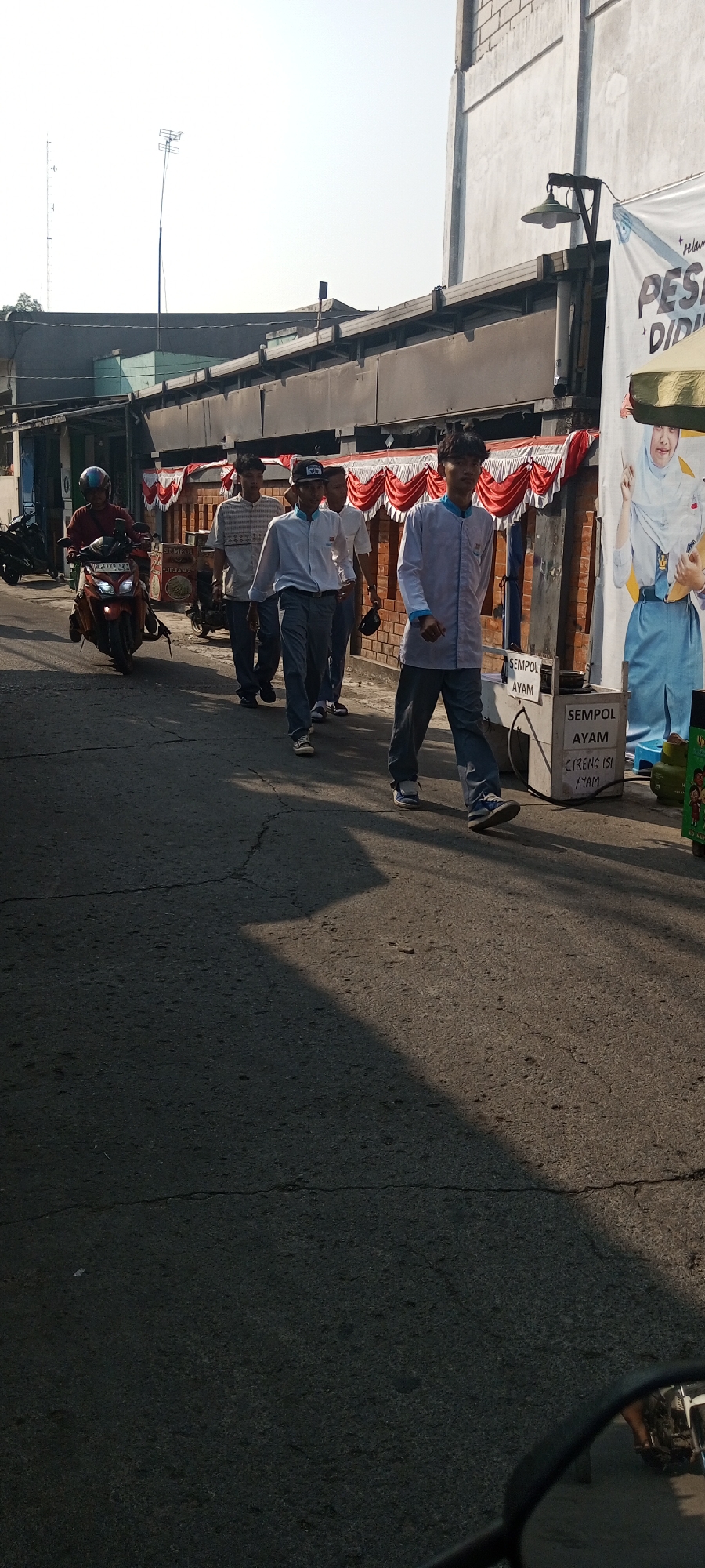 In Setu, Indonesia, on a sunny day, a group of young males in school uniforms are walking down a paved street lined with buildings and decorated with red and white banners. One individual is on a red motorcycle, facing the same direction as the pedestrians. In the foreground, a motorcycle's side mirror reflects the scene. A white stand with text indicating "SEMPOL AYAM" is visible on the right side of the street. The lighting suggests it's late afternoon, casting long shadows across the road.