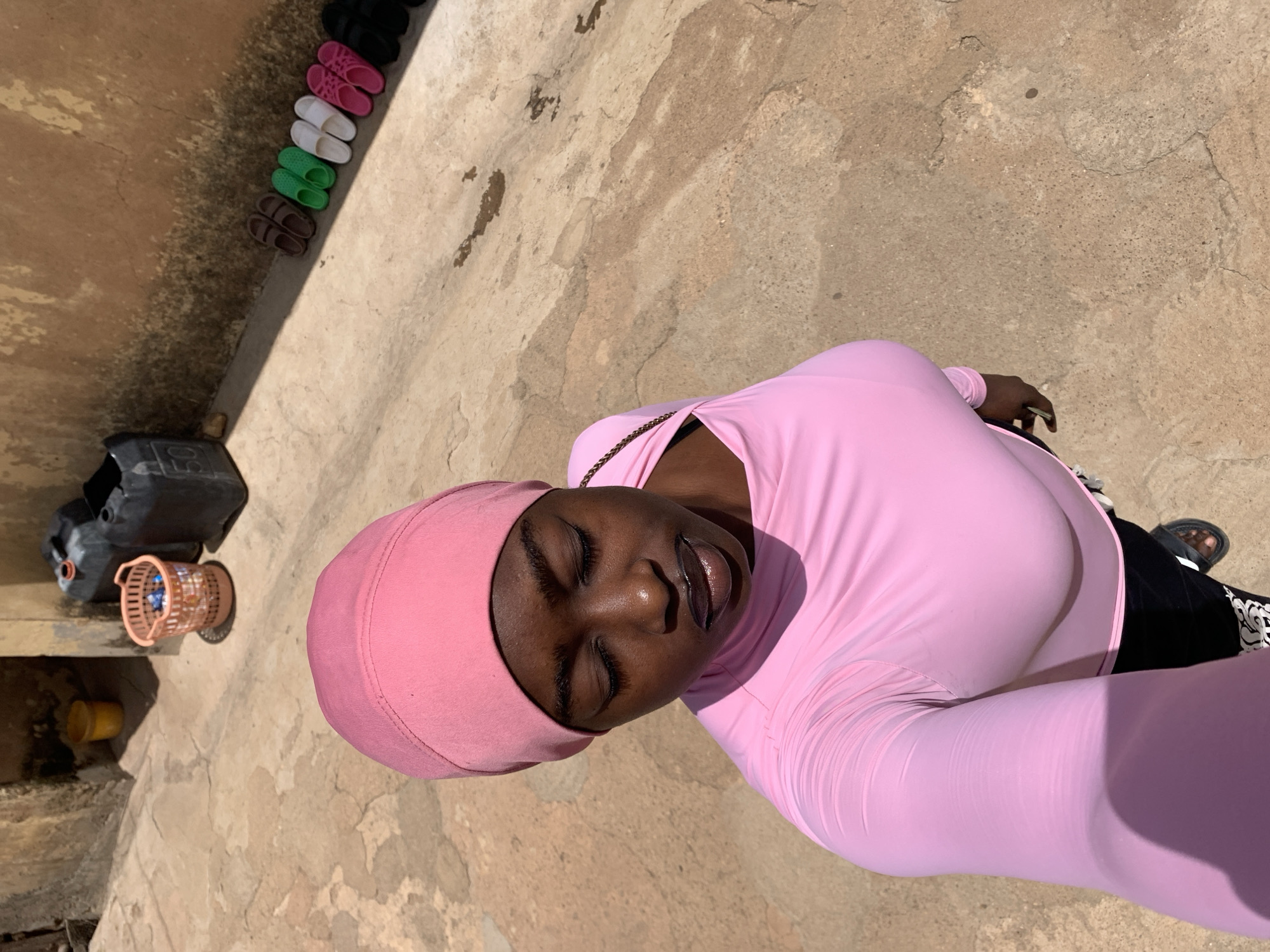 A woman wearing a pink headscarf and a pink, long-sleeved shirt is positioned in the foreground of the image. She is looking directly at the camera with her eyes closed.
To the left of the woman, along a wall, there's a collection of flip-flops of different colors arranged in a row, followed by two black plastic containers, a brown container with blue contents inside, and a small yellow bucket.
The background appears to be a textured, concrete or stone surface. Based on the provided information, the location is Rafin Par, Nigeria.