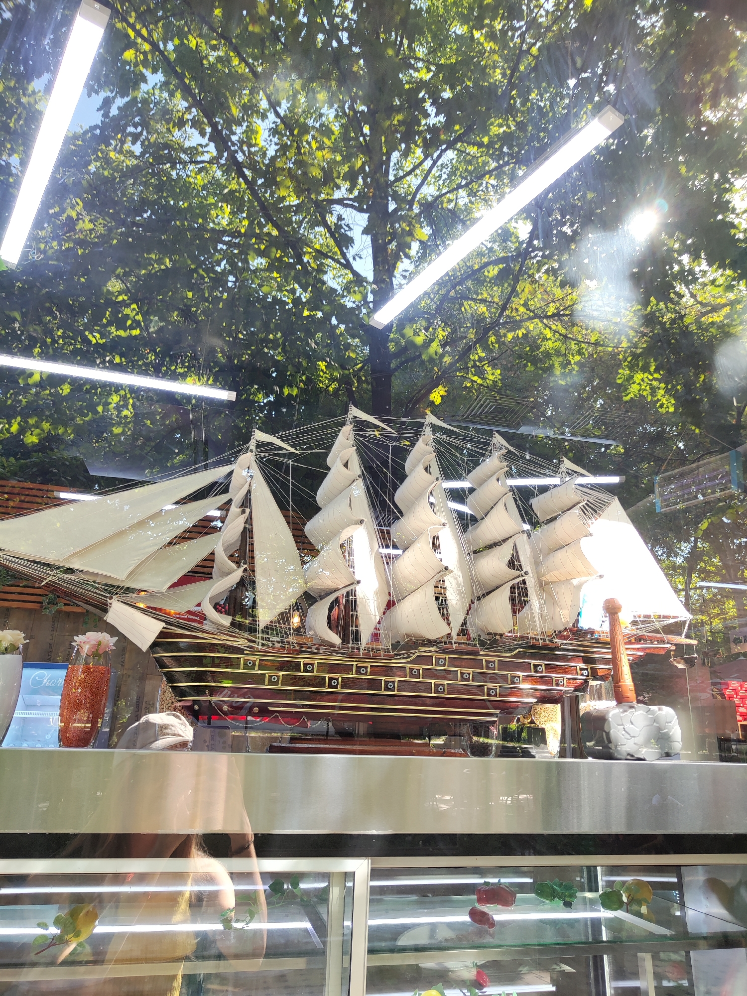 The image shows a model of a large sailing ship with multiple masts and sails, displayed behind glass. The ship is detailed with a dark wooden hull, gold trim, and white sails. Above the ship, there are trees with green leaves and sunlight filtering through, suggesting an outdoor setting or a display near a window. The reflection in the glass shows fluorescent lights and part of what appears to be an interior space with shelves displaying small, colorful objects, possibly candy or sweets. A person's reflection is visible, wearing a hat and a light-colored top, and appearing to be looking down. The scene appears to be in a shop or display area, possibly a confectionary or gift shop. Given the location is specified as Tashkent, Uzbekistan, this could be a display within a commercial establishment in the city.