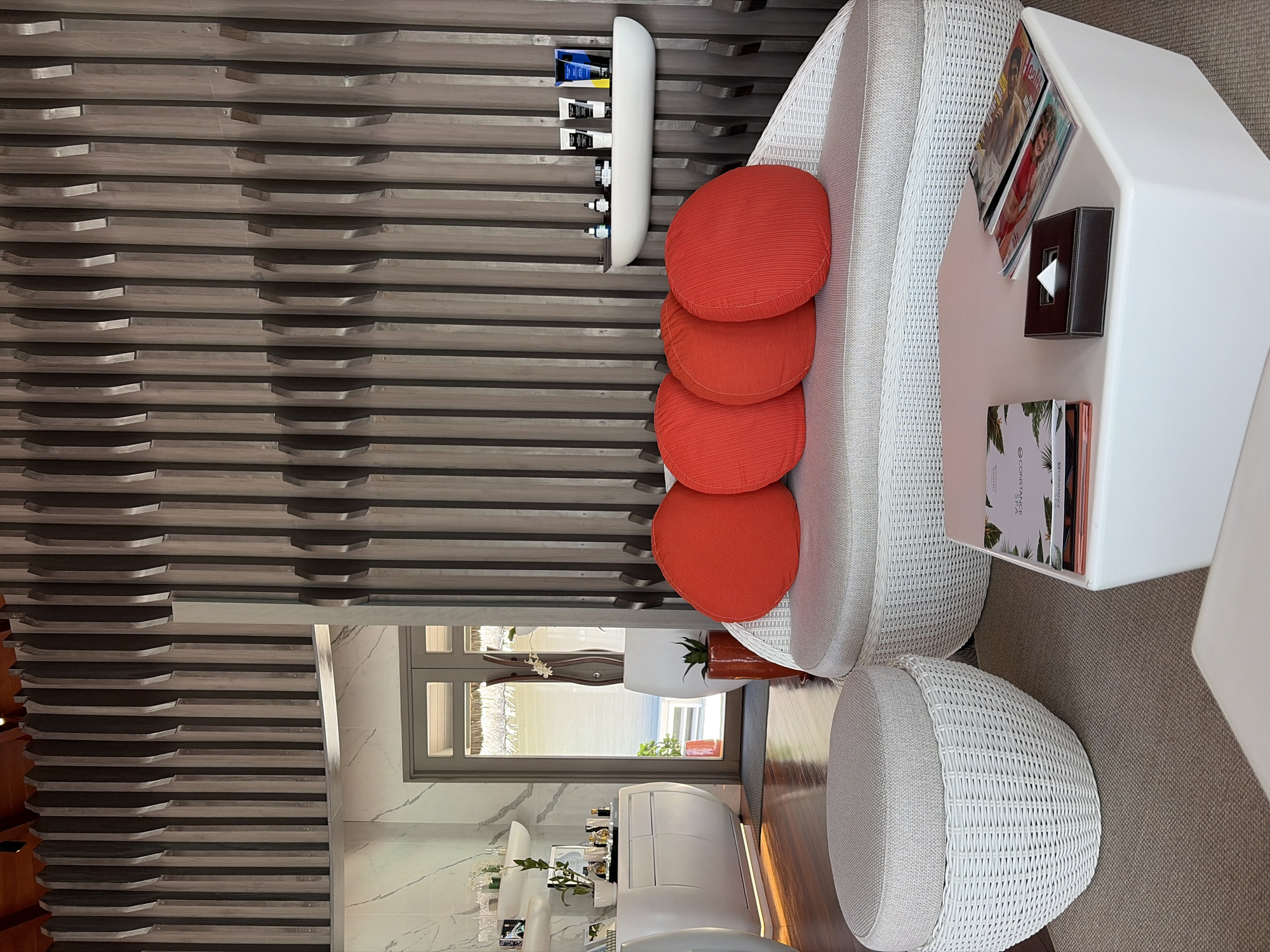 The image depicts a waiting area at a spa in Halaveli, Maldives. The room features a contemporary design with a textured wooden slat wall as a prominent backdrop. In the foreground, a white woven wicker sofa is adorned with four bright red circular cushions. Adjacent to the sofa, a white cube-shaped coffee table holds a dark brown tissue box and several magazines. To the left of the sofa is a round, light gray ottoman with a woven wicker base. In the background, a white curved cabinet is visible, displaying several bottles and containers. To the right of the cabinet, a doorway leads to an outdoor area with thatched roofs. The overall ambiance is serene and luxurious, consistent with a high-end spa setting.