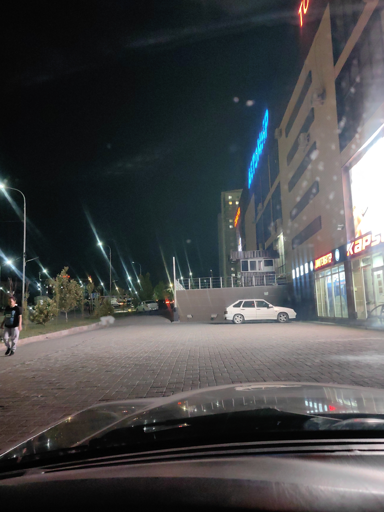An image taken at night shows a street scene in Shymkent, Kazakhstan, possibly near a commercial building. A person in a dark t-shirt and light-colored pants walks on the left side of the paved walkway. A white car is parked on the right side of the street, near a building with illuminated signs. The building appears to be a shopping center or complex, with bright neon lights and large windows. Streetlights are visible along the road, casting long reflections on the wet pavement. The overall ambiance is nocturnal, with artificial lights dominating the scene.