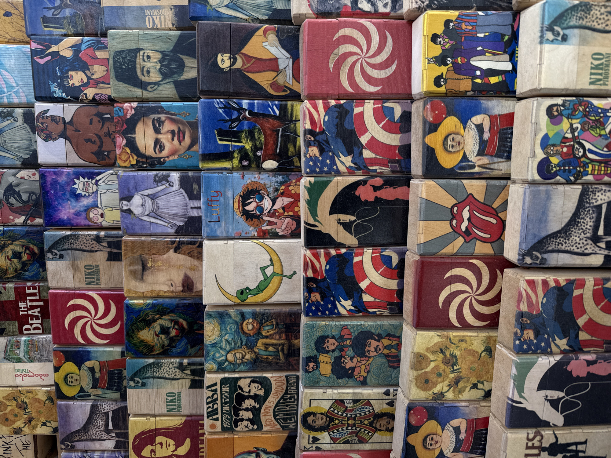 The image presents a close-up view of a stacked arrangement of small, rectangular wooden boxes. Each box features a unique design or illustration on its top surface. Some of the designs include portraits of famous figures like Frida Kahlo and Tupac Shakur, pop culture references such as characters from the animated series Rick and Morty and One Piece, artwork inspired by The Beatles and Van Gogh, and logos like The Rolling Stones' iconic tongue symbol. The boxes are arranged in rows and columns, creating a visually dense mosaic of images. Words are visible on some boxes including "NIK" "ABBA", "THE BEATLES" and "PIROSMANI". The image shows a collection of souvenir-type boxes.
The location in the city of Mtskheta, Georgia is unknown.