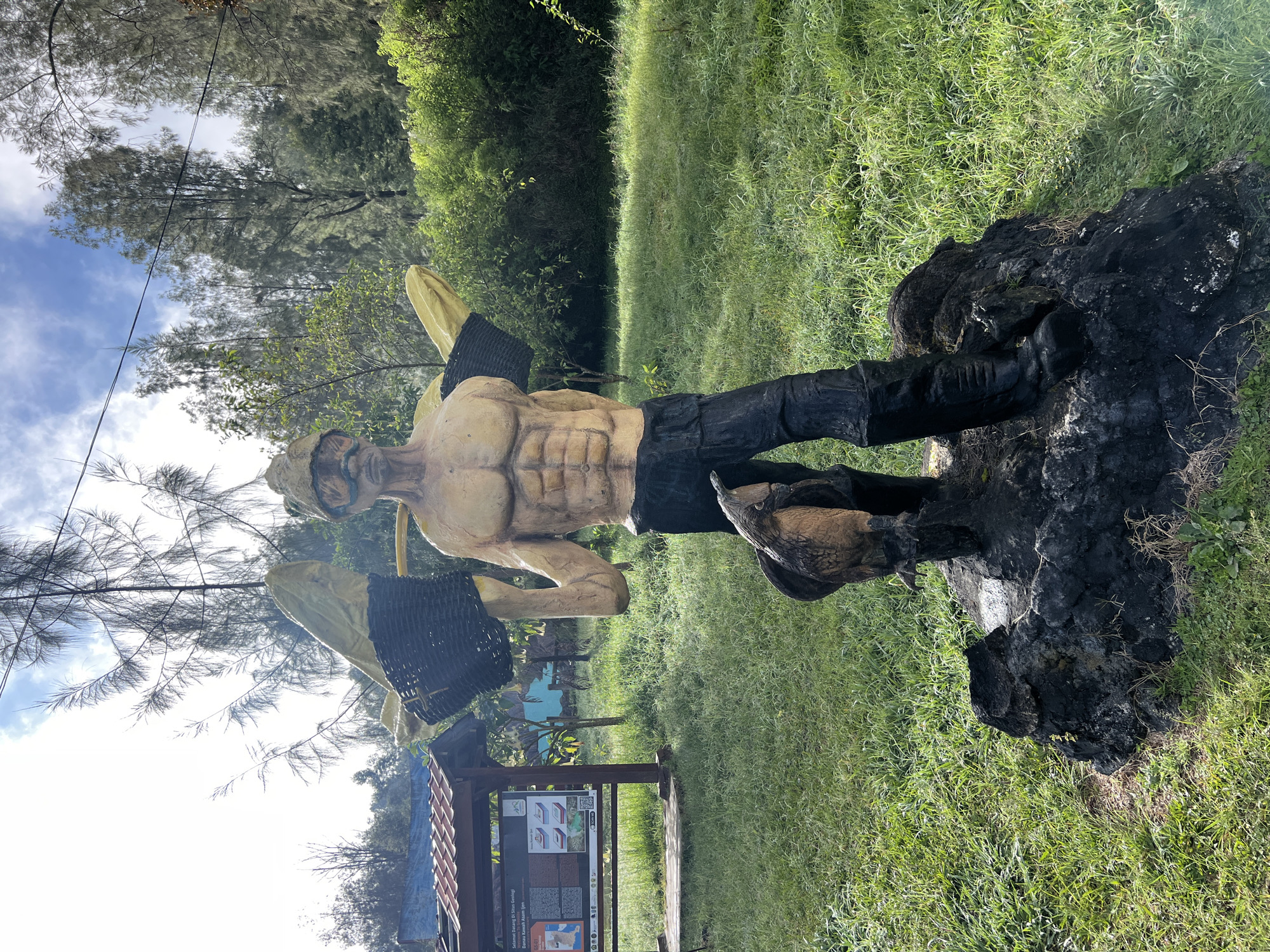 The image captures a statue of a muscular male figure wearing what appears to be traditional Indonesian attire, including headwear and a sarong. The statue is positioned on a dark, possibly volcanic rock formation amidst a grassy, green landscape. The setting is outdoors, suggesting a park or garden area. In the background, there is dense vegetation and trees, with glimpses of the sky through the branches. To the left of the statue, there is a wooden information board under a small shelter, indicating a tourist or informational site. The location is in the city of Unknown, Indonesia.